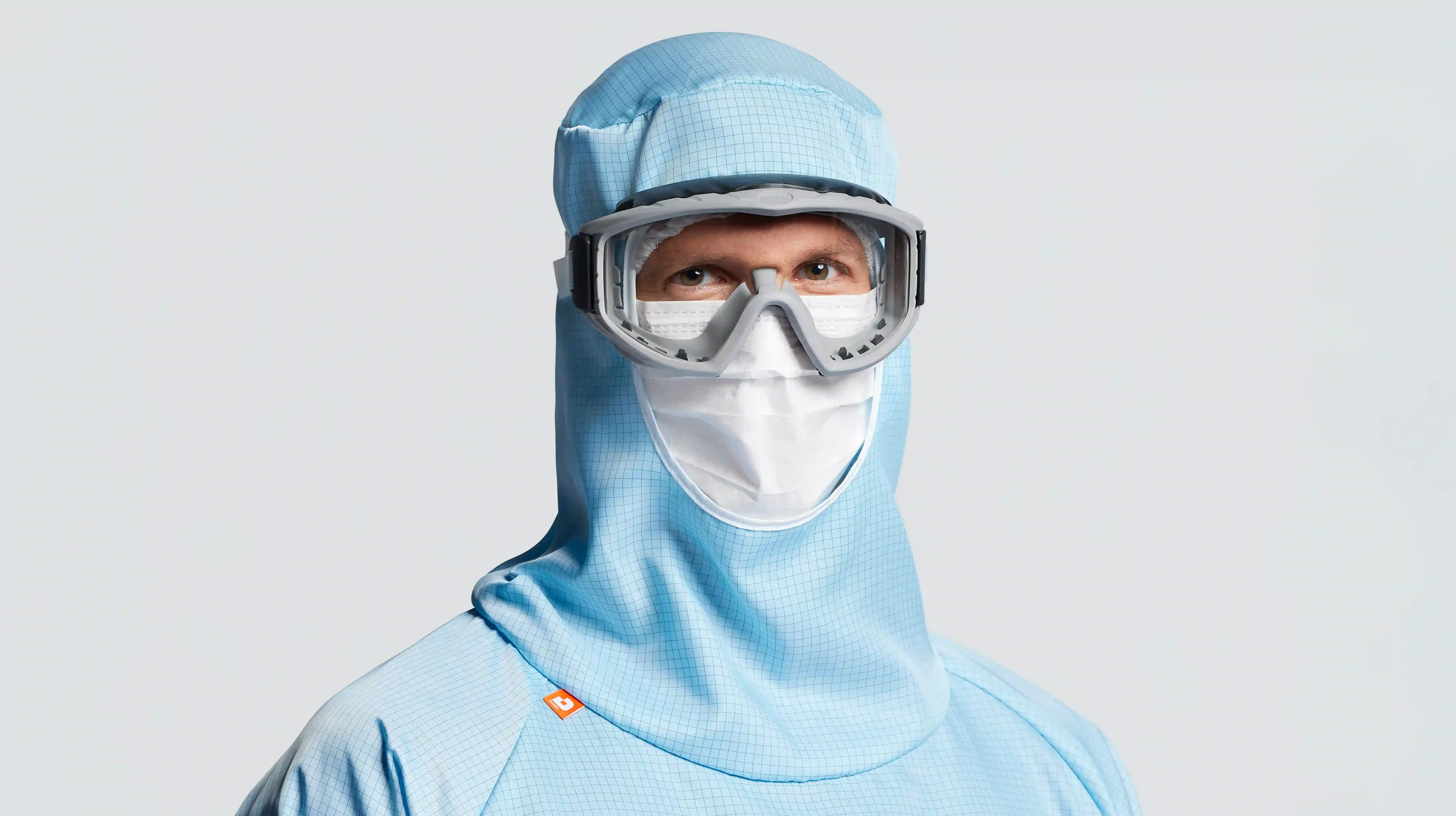 Person in blauer Schutzkleidung mit Haube, Schutzbrille und Gesichtsmaske vor hellem Hintergrund.