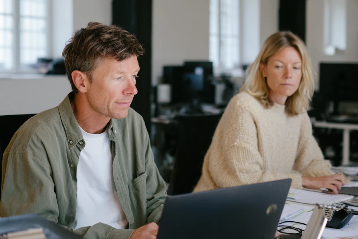 Two coworkers seated side by side working on laptops in an office with bright windows in the background.