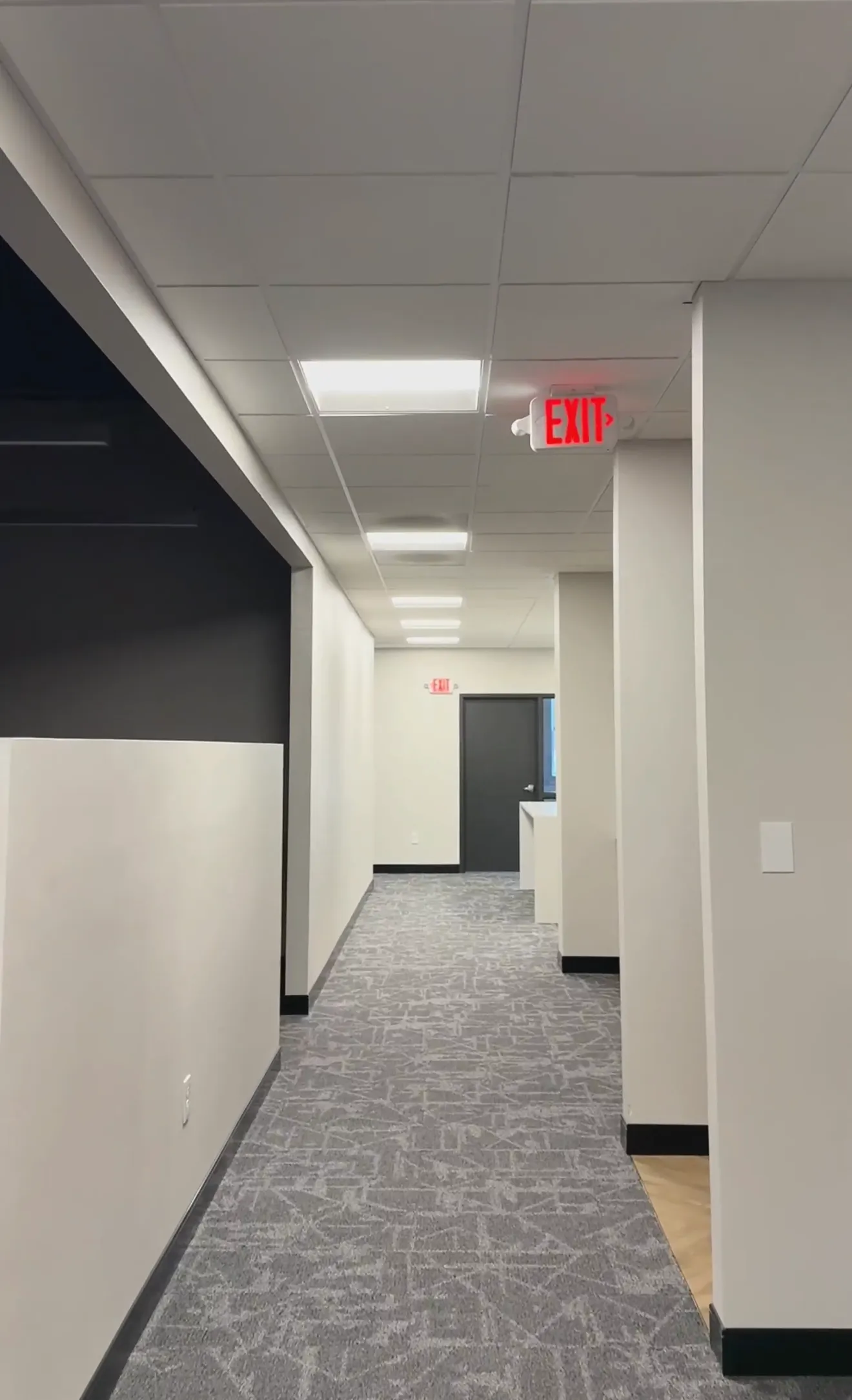 Empty office hallway with white walls, gray carpet, and red exit sign
