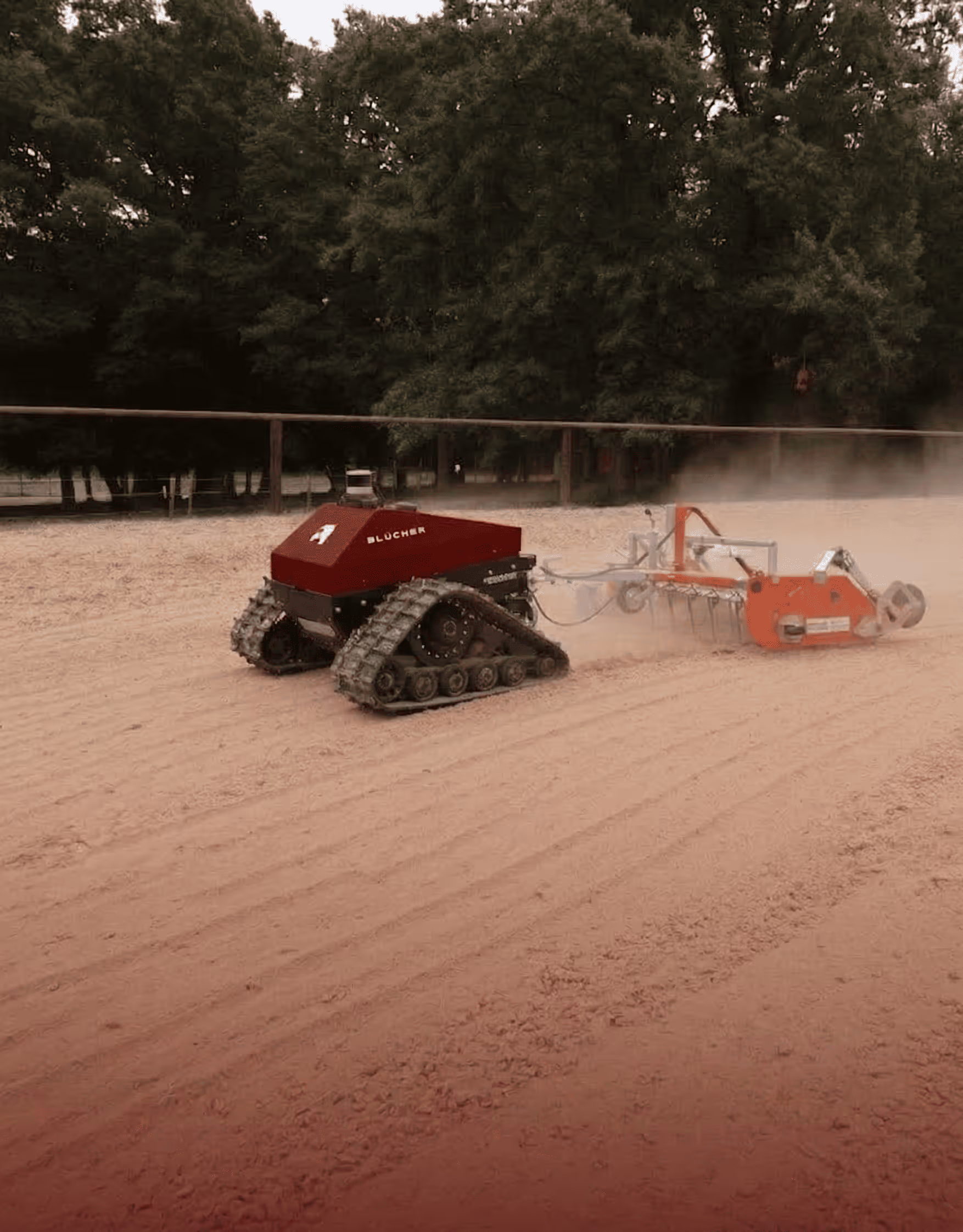 Roter Blücher-Roboter mit Panzerspuren, der einen sandigen, von Bäumen umgebenen Reitplatz pflegt.