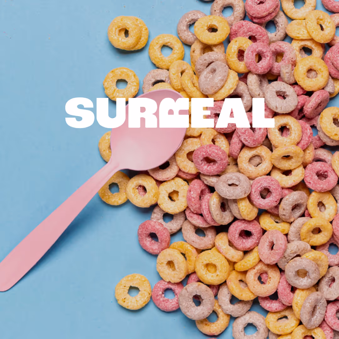 Pink spoon on blue surface surrounded by yellow, pink, and purple ring-shaped cereal with the word SURRÆAL in white text.