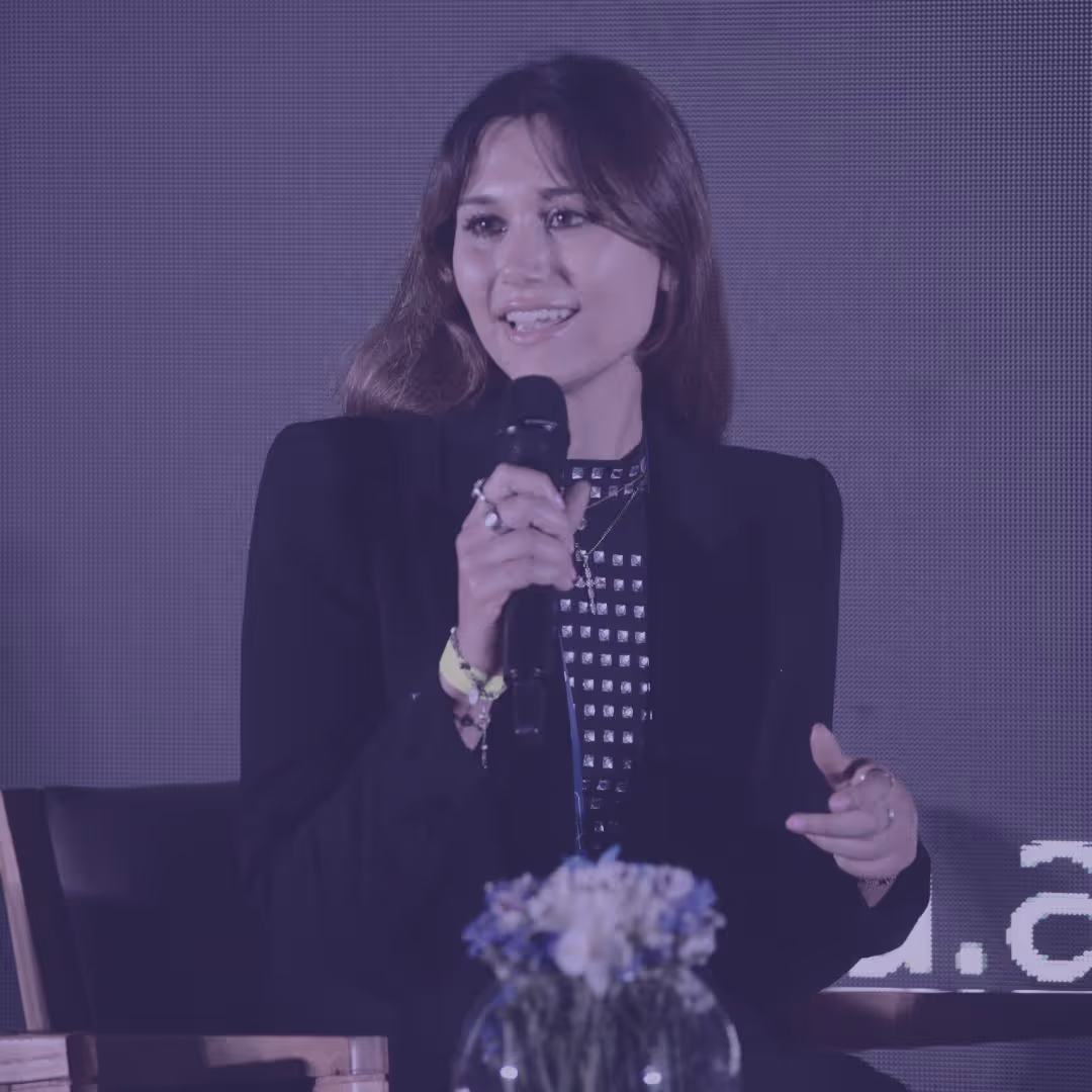 Woman speaking into a microphone during an event, wearing a black blazer and patterned top, with a flower arrangement in front.