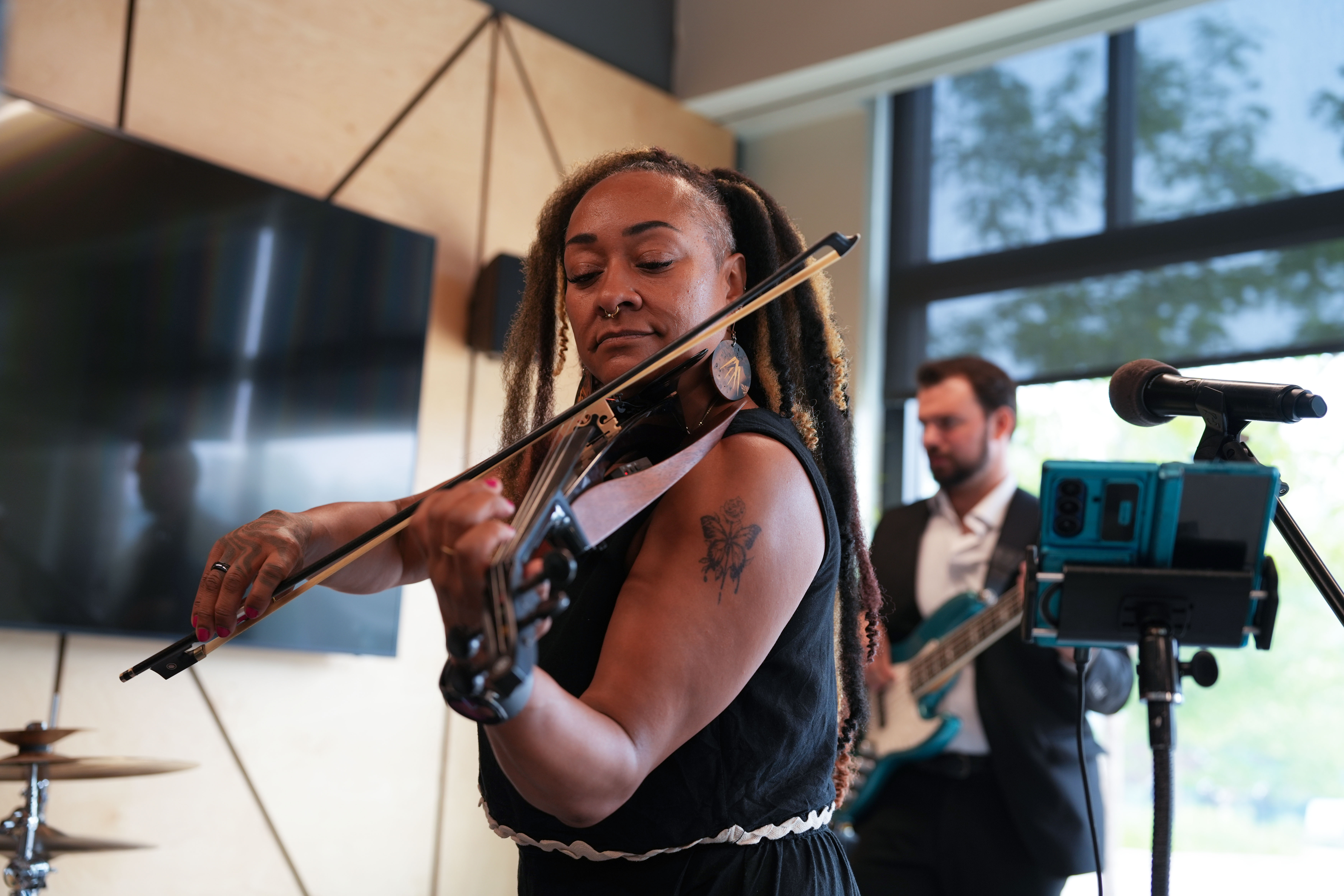 Musician playing five-string electric violin live indoors with a background bass guitar player and microphone setup.