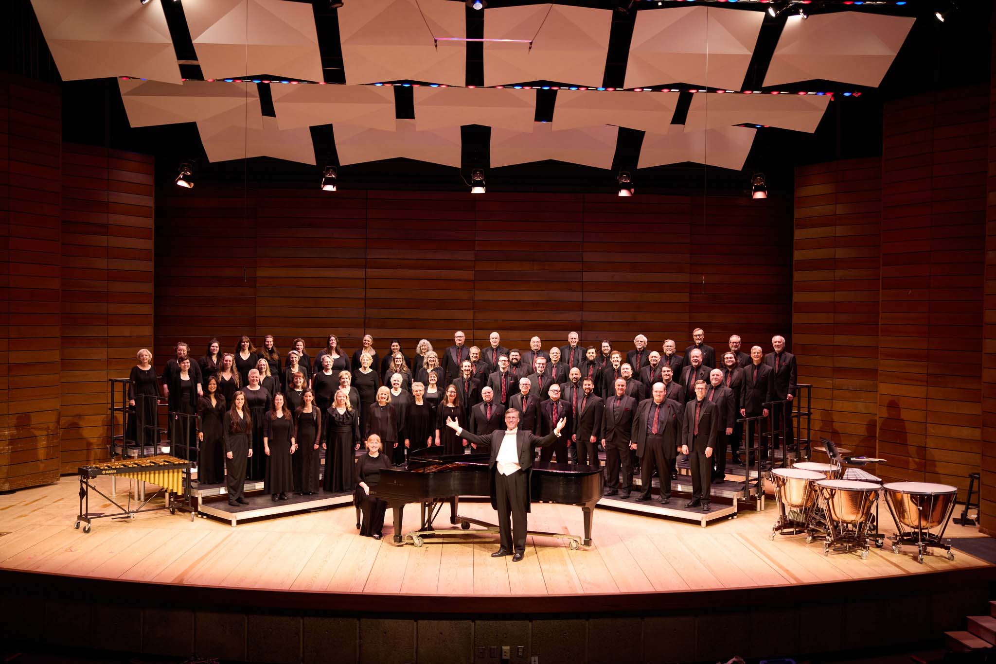 Large choir choir in dark attire standing on stage with a conductor in a white-tie tuxedo acknowledging the audience next to a grand piano.