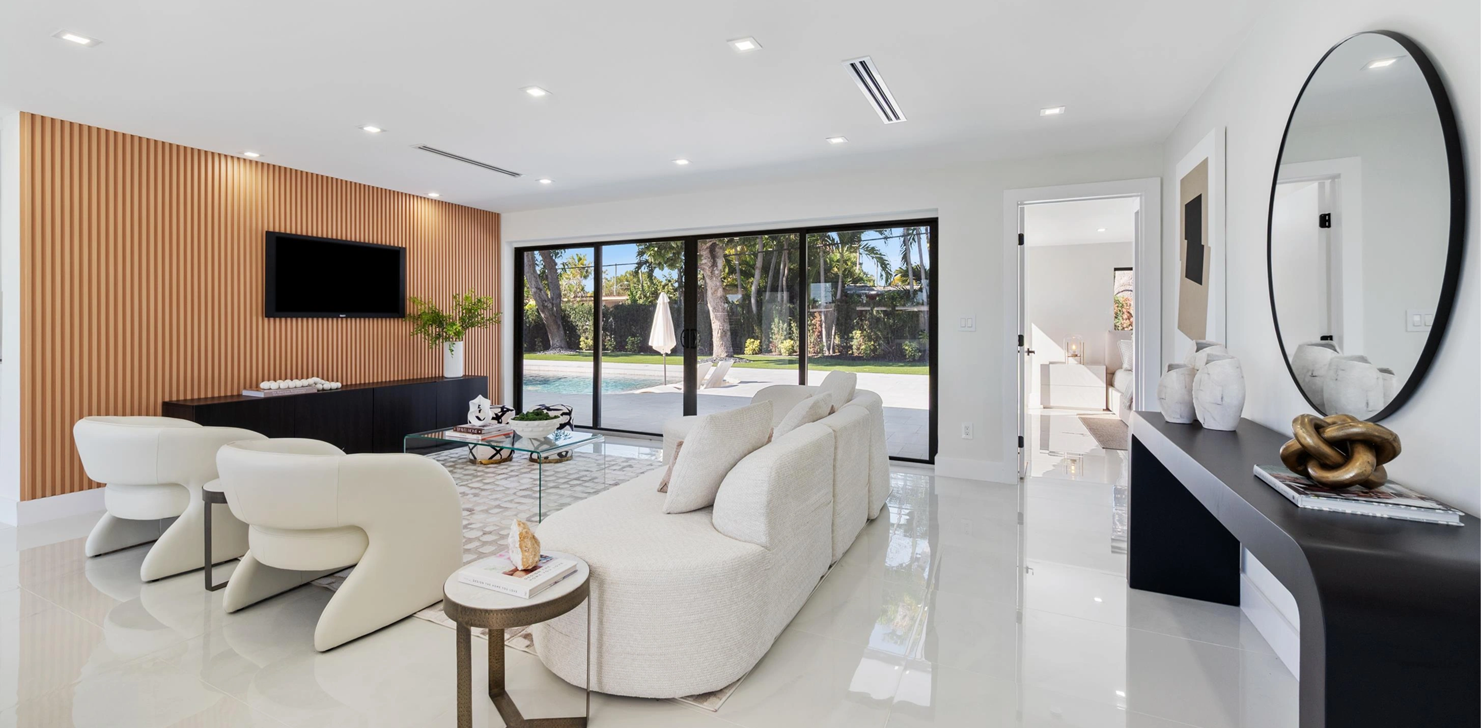 Modern living room with glass coffee table, curved white chairs, sofa, wood slat TV wall, and patio view.