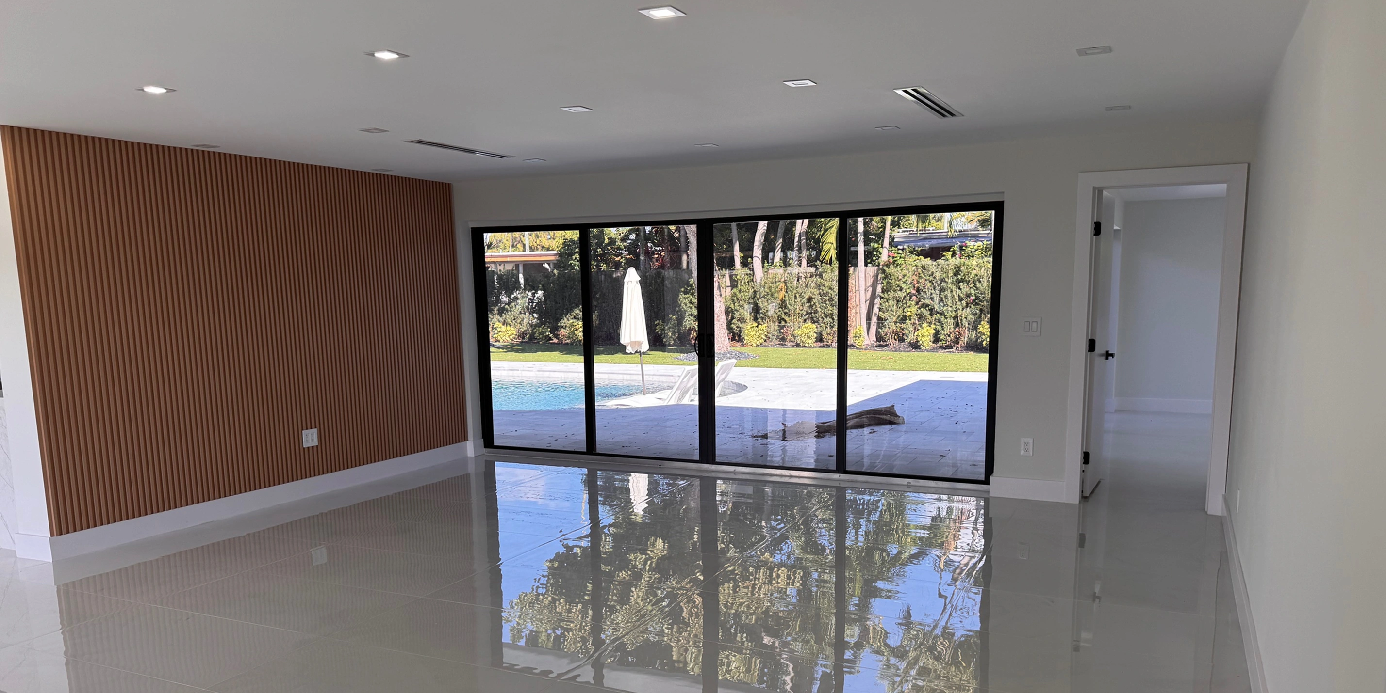 Empty modern room with glossy floor, wood-paneled wall, sliding glass doors opening to poolside patio.