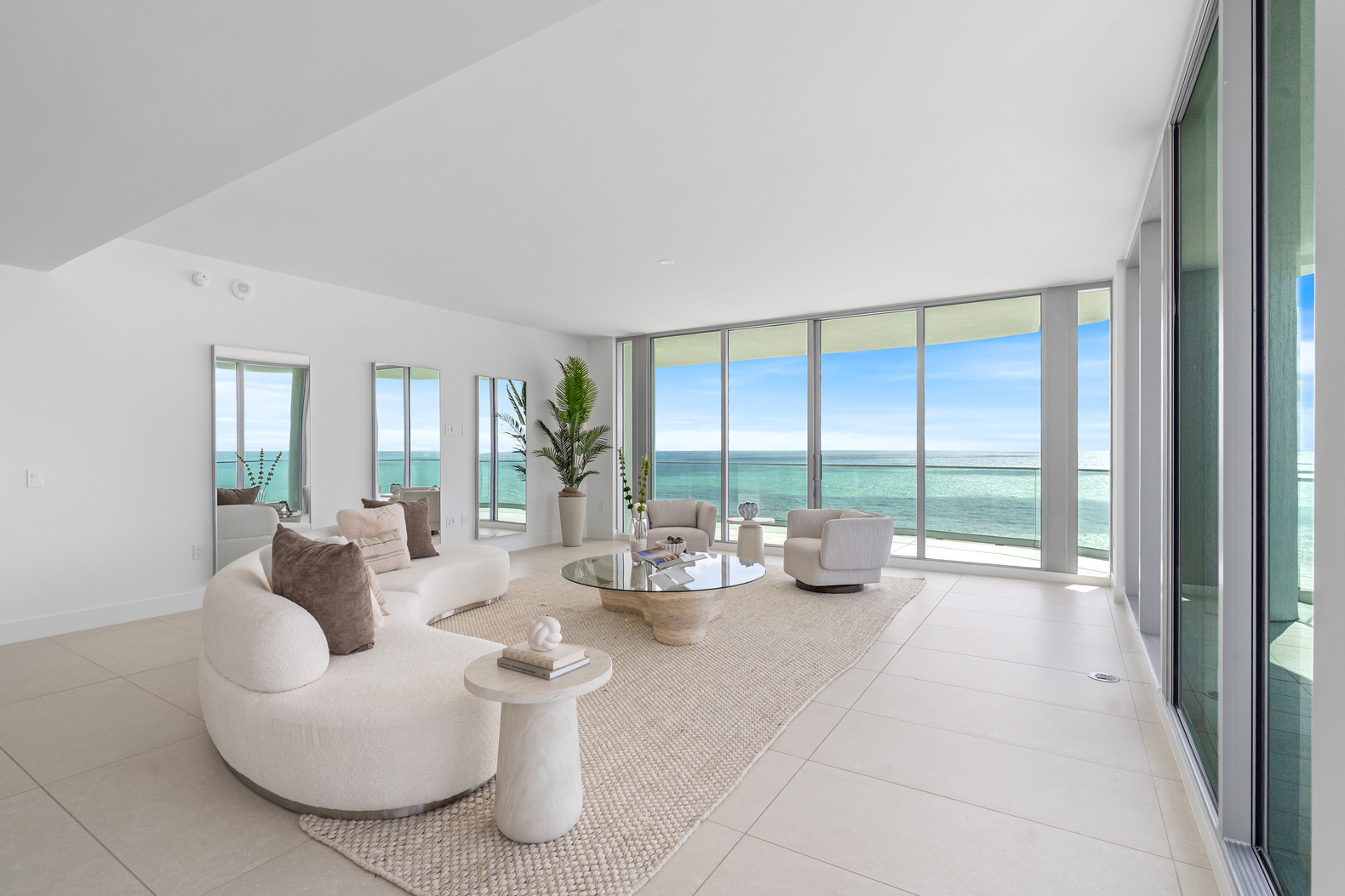 Bright living room with curved white sofa, armchairs, glass coffee table, large windows overlooking ocean.