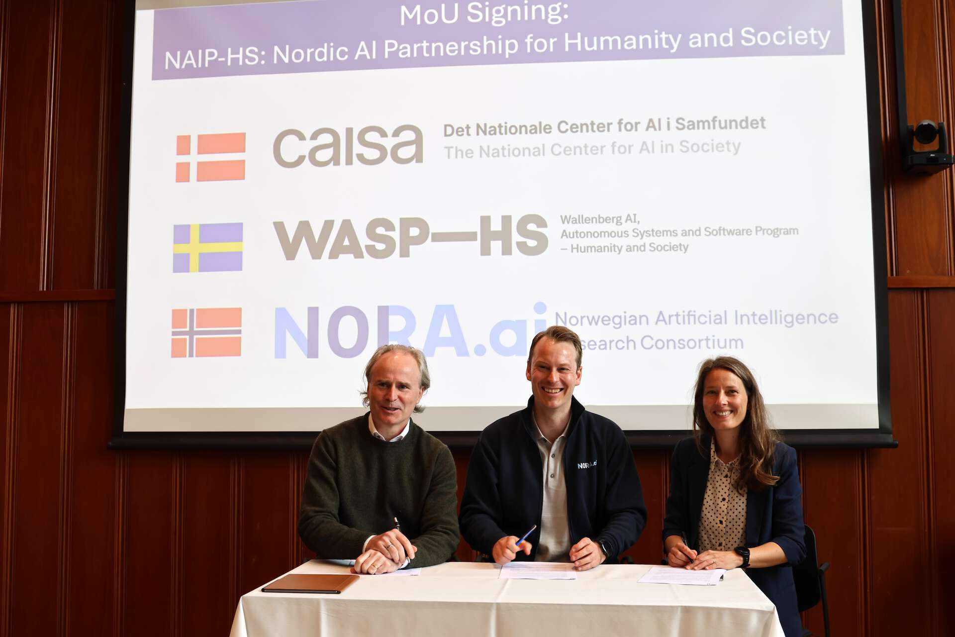 Three people sitting by a table, smiling towards the camera. In the background, a screen with logos of CAISA, WASP-HS and NORA