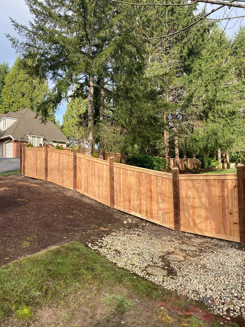 Newly built cedar privacy fence with dark posts and caps following a sloped yard, bordering a wooded residential property