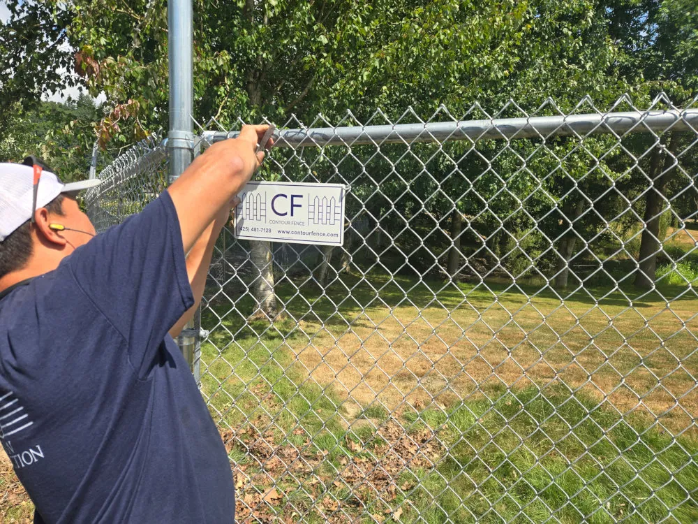 Contour Fence technician attaching a CF company sign to a galvanized chain-link fence, wearing safety gear, with grassy park and trees in the background