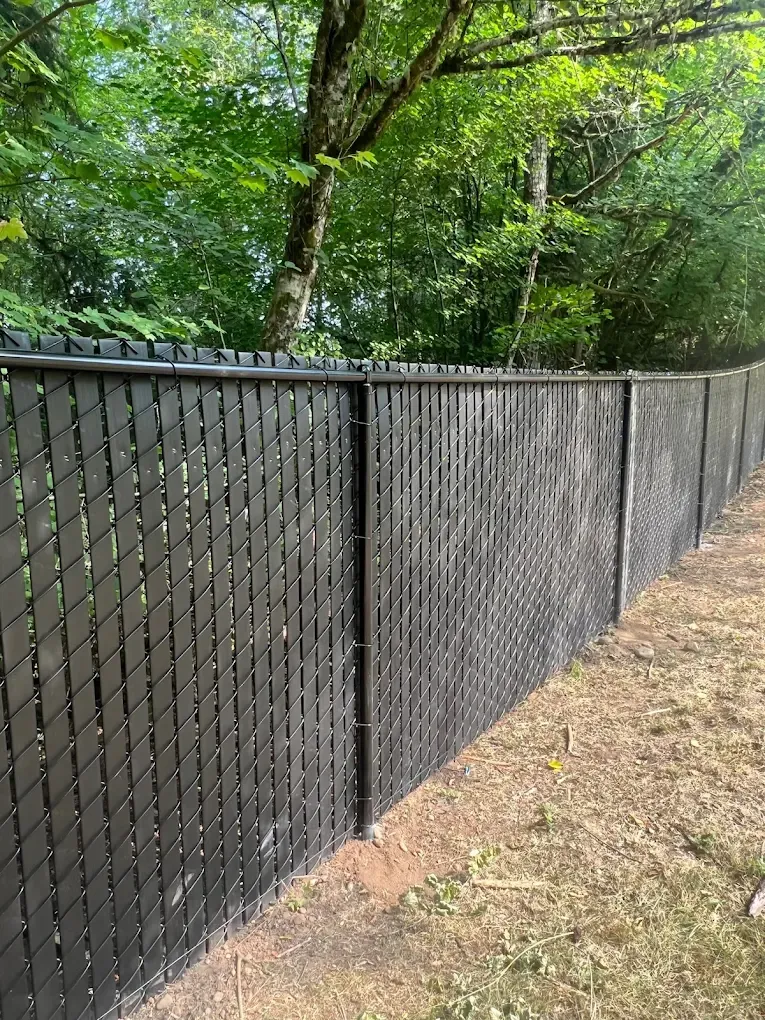 Black chain link fence equipped with vertical privacy slats along a wooded tree line.