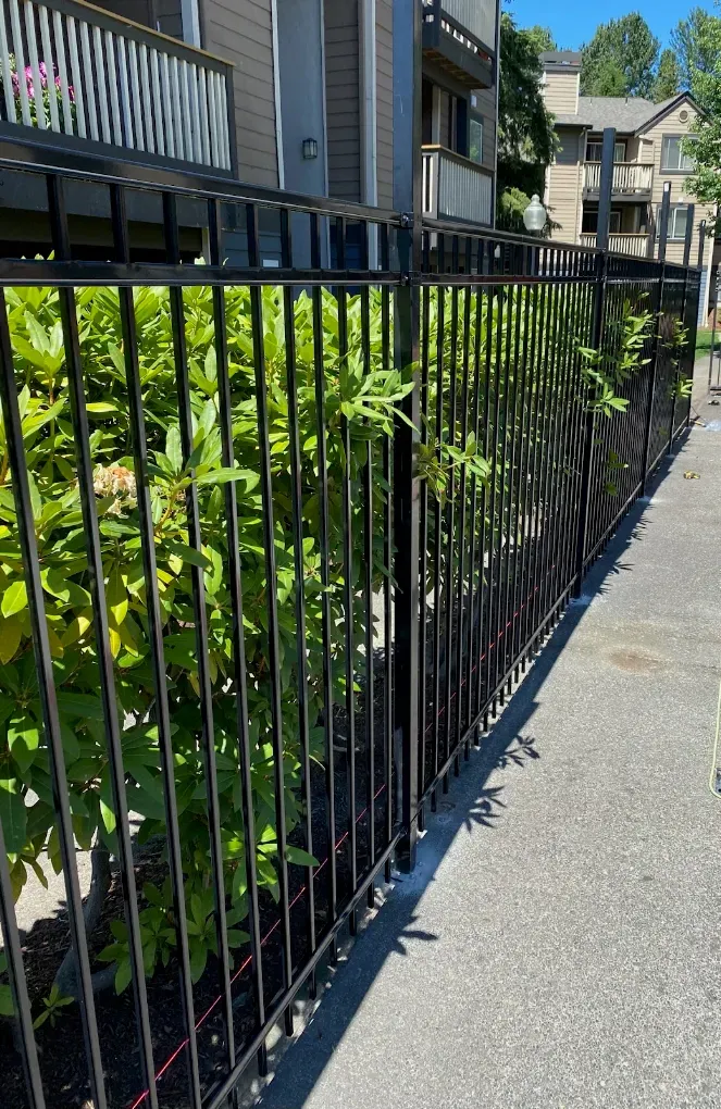 Black metal security fence installed alongside leafy green hedges and a paved walkway.
