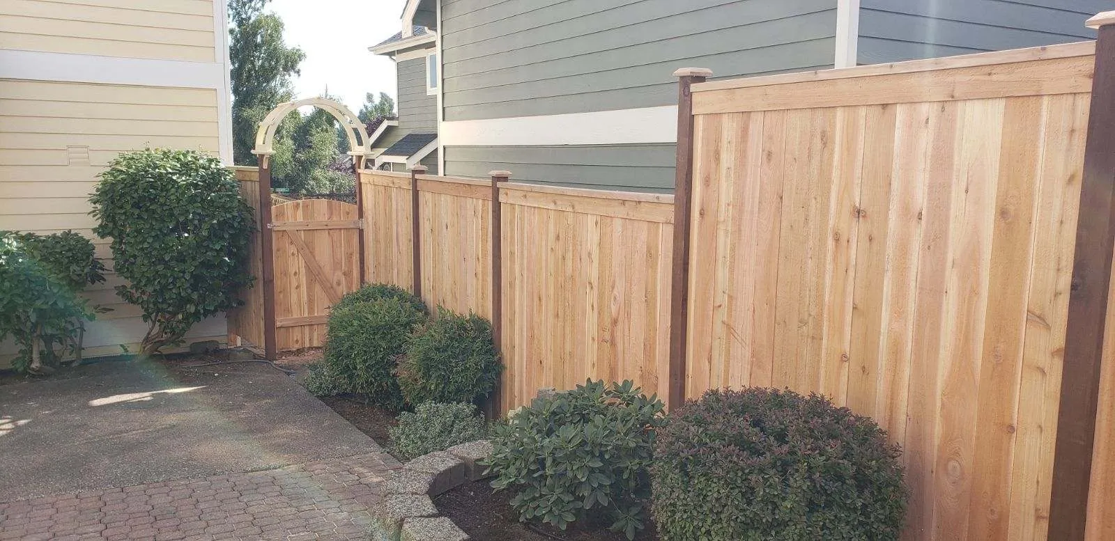 Natural wood privacy fence leading to an arched garden gate next to a house exterior and shrubs.