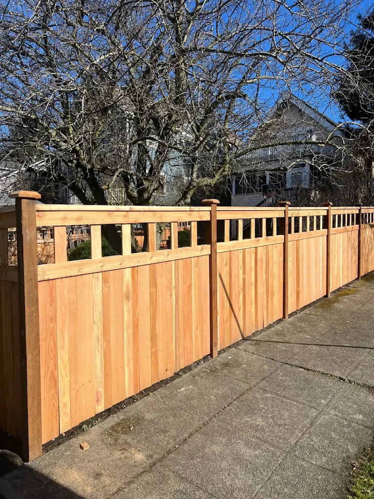 Natural cedar privacy fence with open vertical slat top trim running along a concrete sidewalk.
