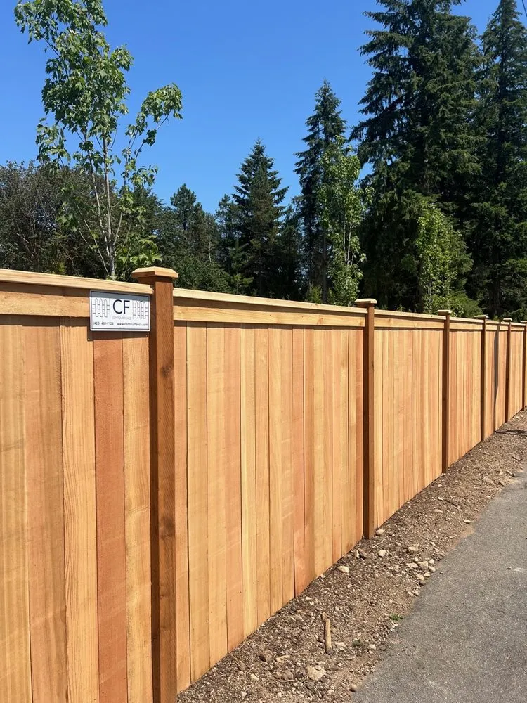 Tall natural wood privacy fence with post caps installed along a paved walkway.