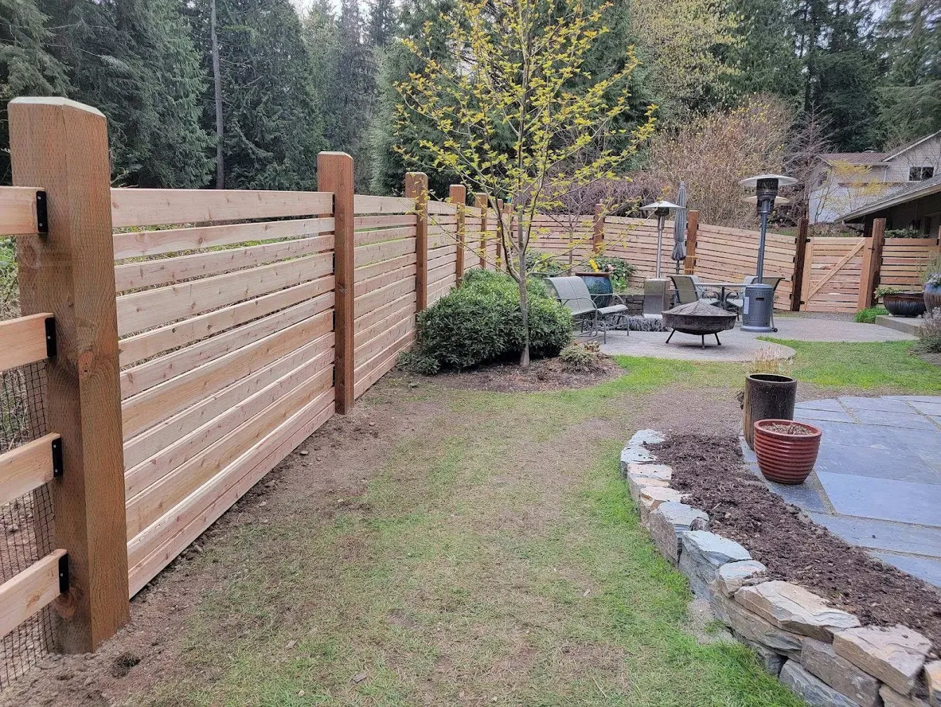 Horizontal slat wooden fence with black metal mounting brackets enclosing a patio area with a fire pit.