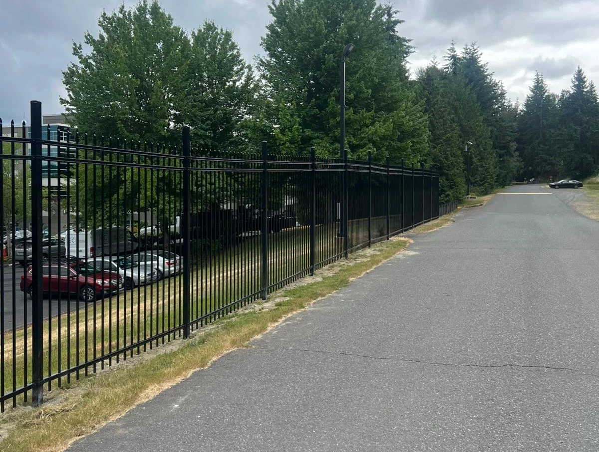 Black wrought iron perimeter security fence installed along a commercial paved driveway with green trees in the background.
