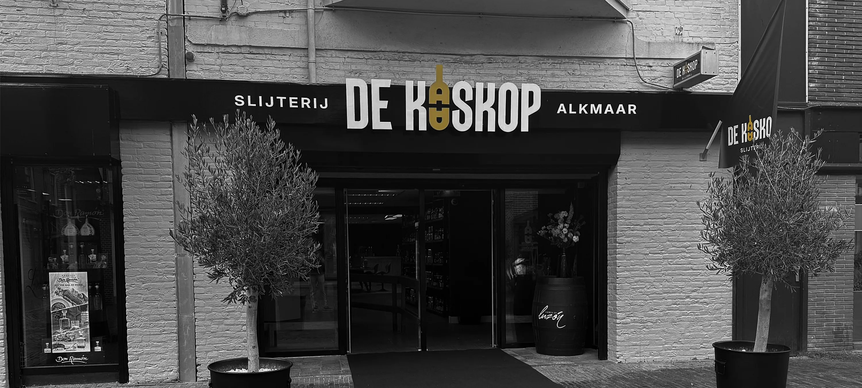 Zwart-wit gevelaanzicht van slijterij De Kaaskop in Alkmaar, met het logo boven de ingang, twee olijfbomen in potten en een wijnvat bij de entree.