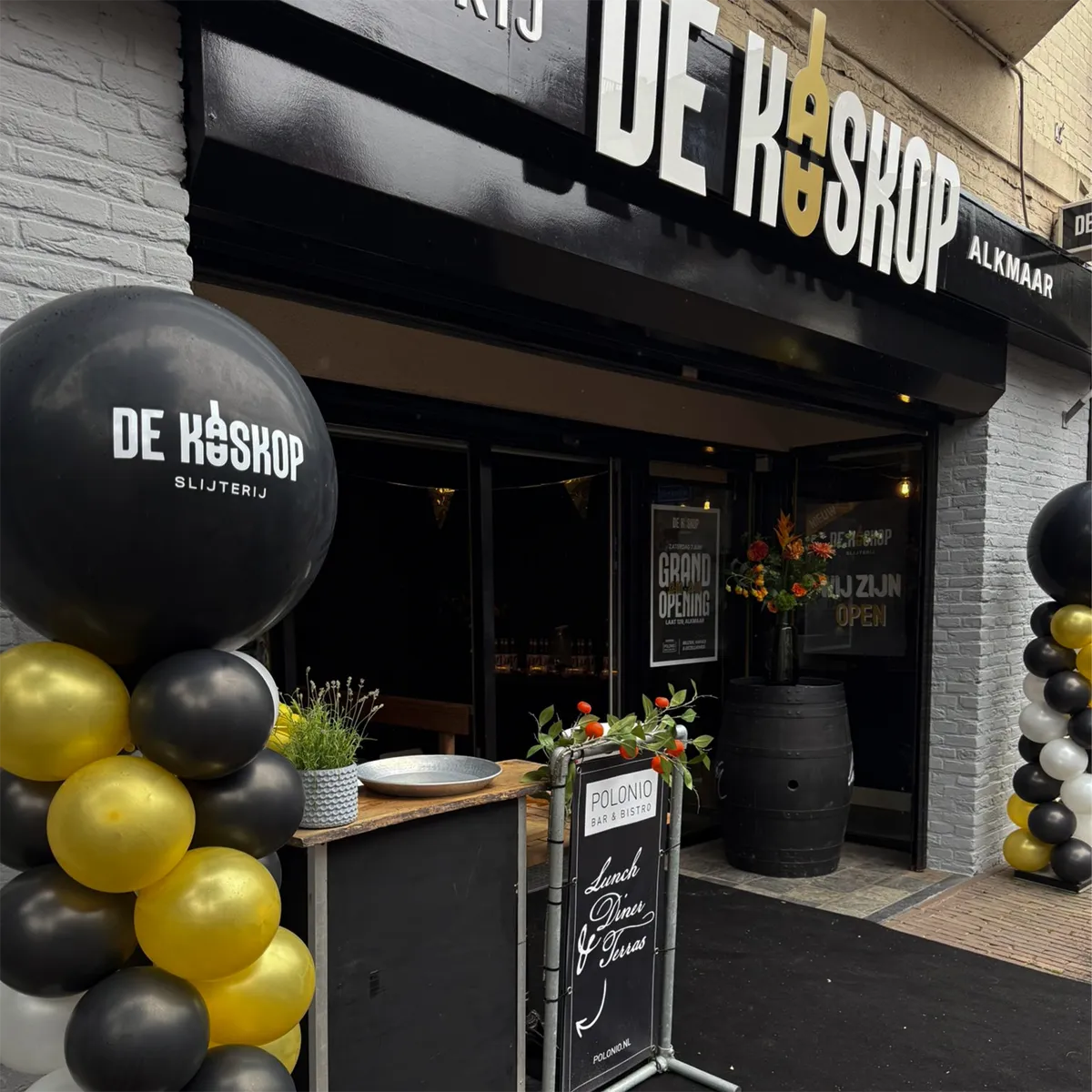 Entree van slijterij De Kaaskop in Alkmaar tijdens de opening, met ballonnen in zwart, goud en wit, een wijnvat met bloemen en een bord met de tekst ‘Grand Opening’.