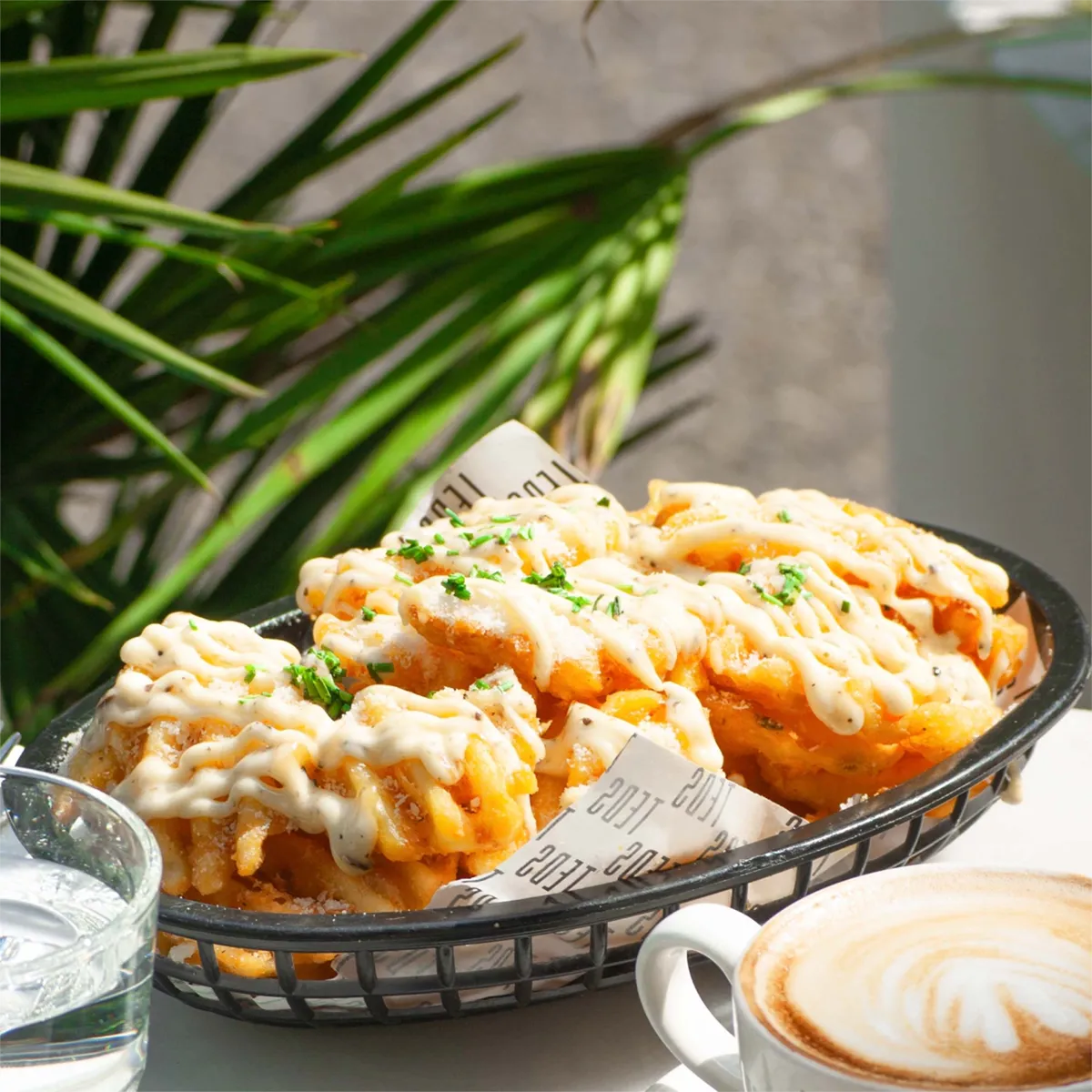 Verse loaded fries met kaassaus en kruiden geserveerd bij TEDS brunch café, naast cappuccino met latte art op zonnig terras.