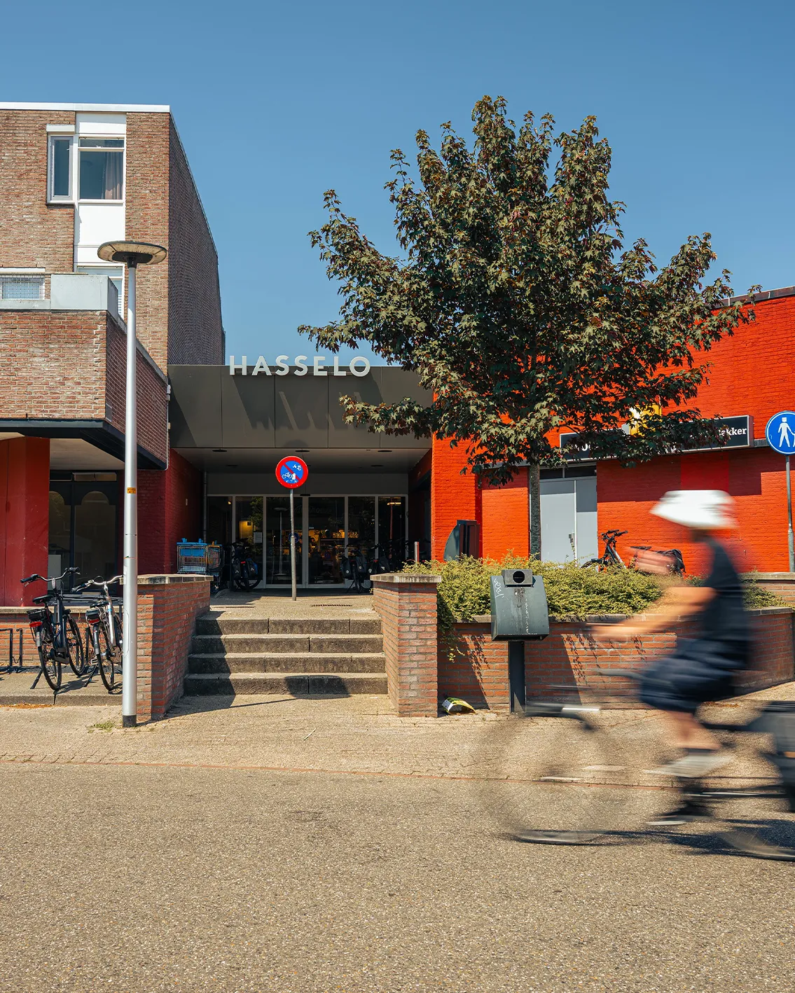 Ingang van Winkelcentrum Hasselo met het HASSELO-logo op de gevel, een oranje pand en een voorbijganger op de fiets.
