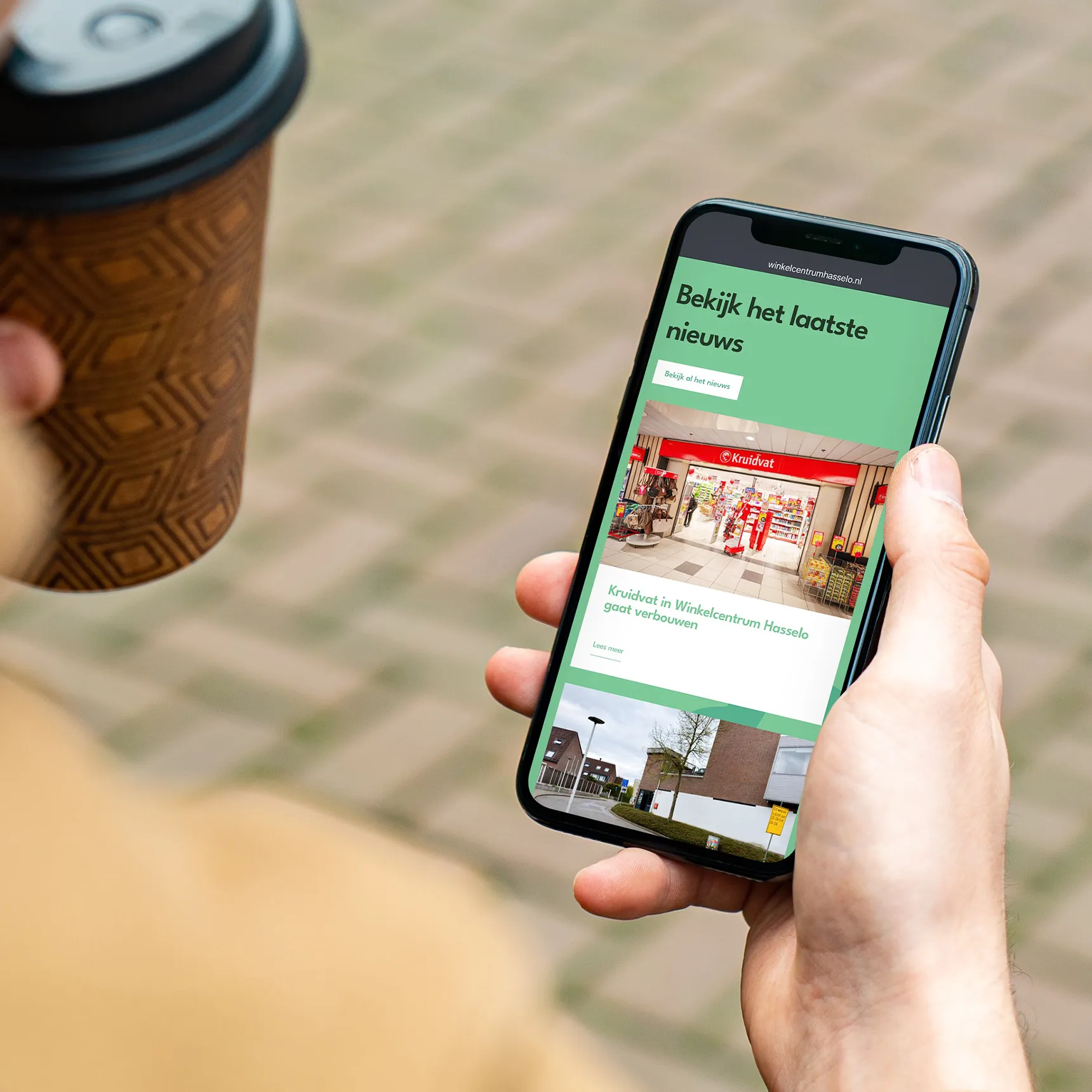 Hand met koffiebeker houdt een smartphone vast met de nieuwspagina van de website van Winkelcentrum Hasselo.
