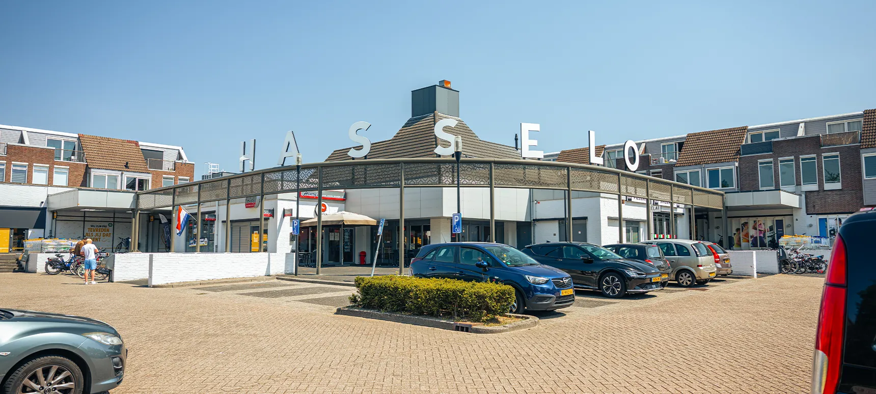 Buitenaanzicht van Winkelcentrum Hasselo met de grote losse HASSELO-letters op het dak en een parkeerplaats op de voorgrond.