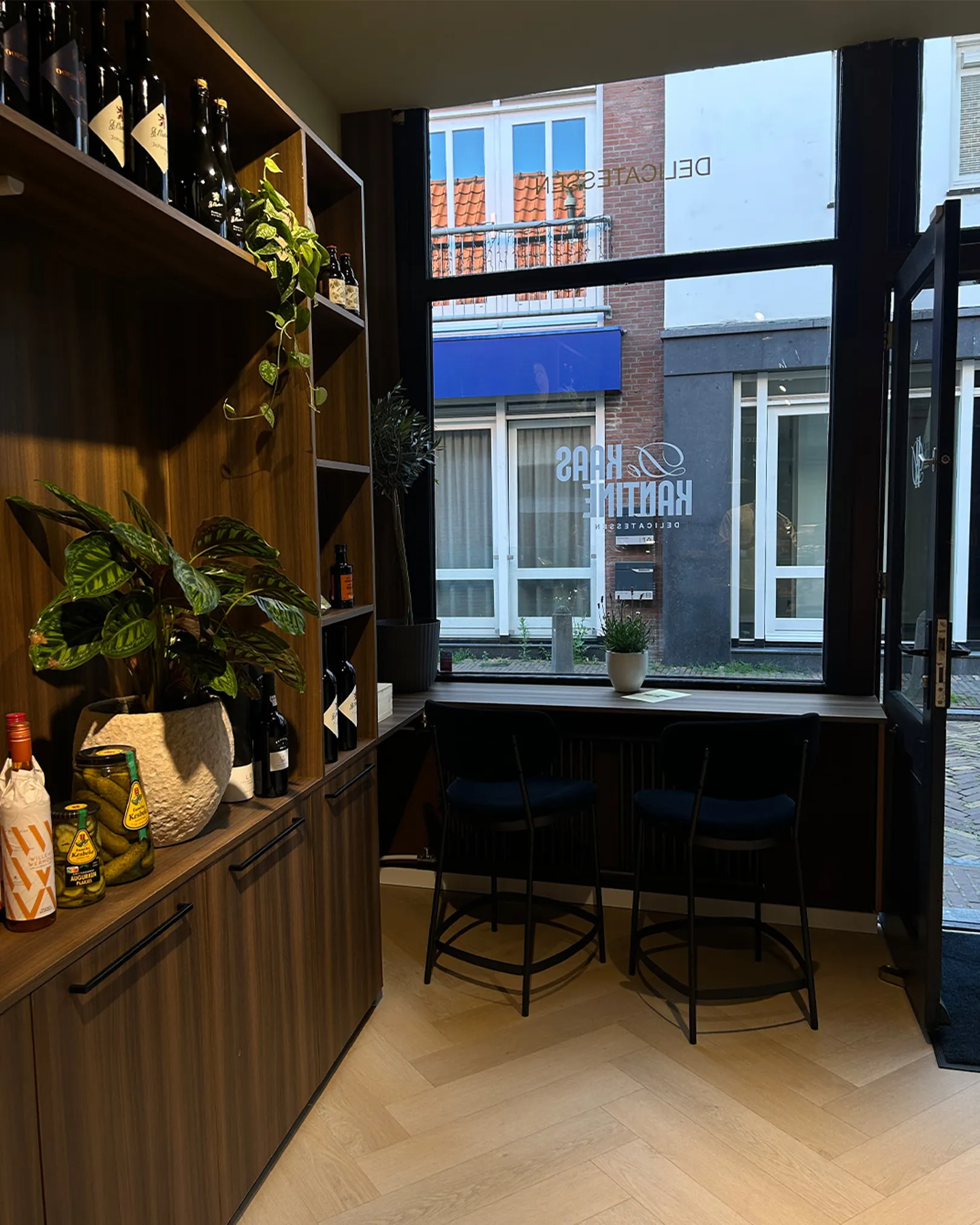 Zithoek bij het raam in De Kaaskantine met barkrukken, planten en de spiegelbeeldige gevelbelettering zichtbaar door het glas.