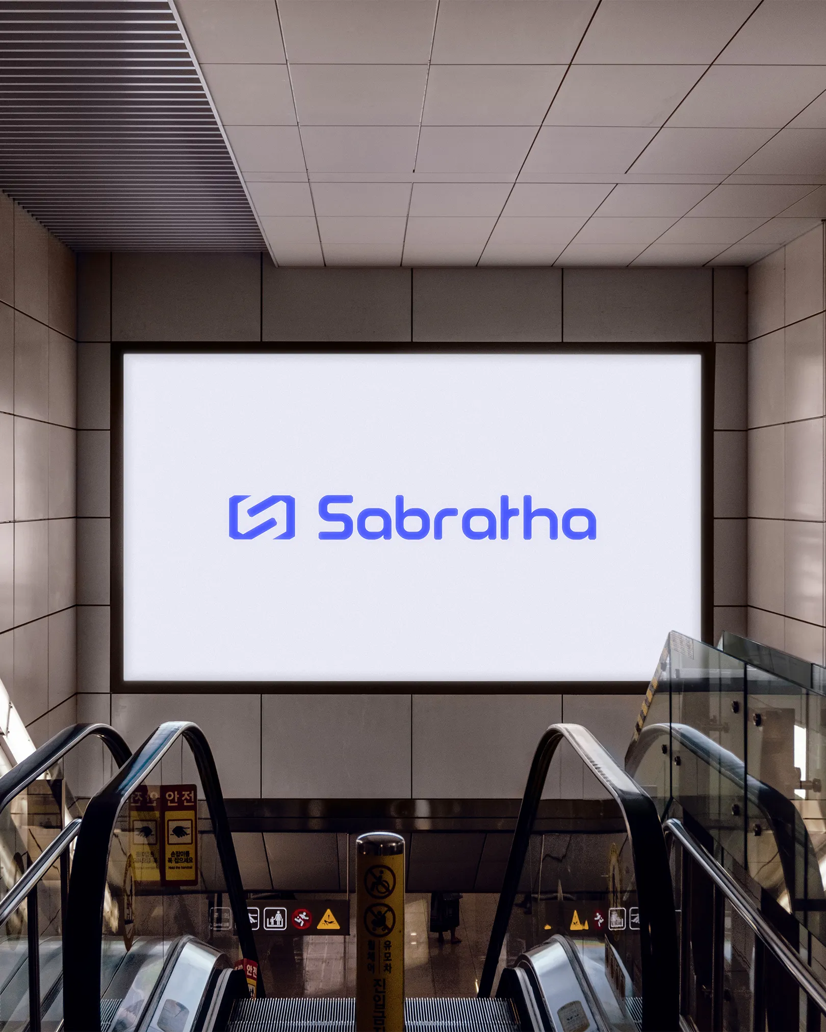 Grote verlichte reclamewand in een moderne metro- of stationsomgeving, boven roltrappen. Op het witte scherm staat het blauwe logo en woordmerk “Sabratha”, strak en centraal gepositioneerd binnen een architectonische ruimte van staal en beton.