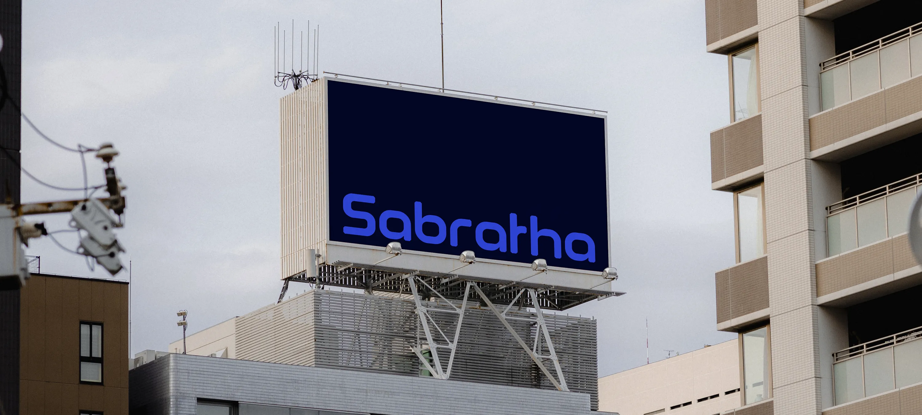 Groot rechthoekig billboard op een stadsdak met een donkerblauwe achtergrond en het woord “Sabratha” in helder blauw. Het bord steekt boven moderne appartementsgebouwen uit en wordt ondersteund door een metalen constructie, wat het merk een krachtige, schaalbare en technologisch georiënteerde uitstraling geeft.