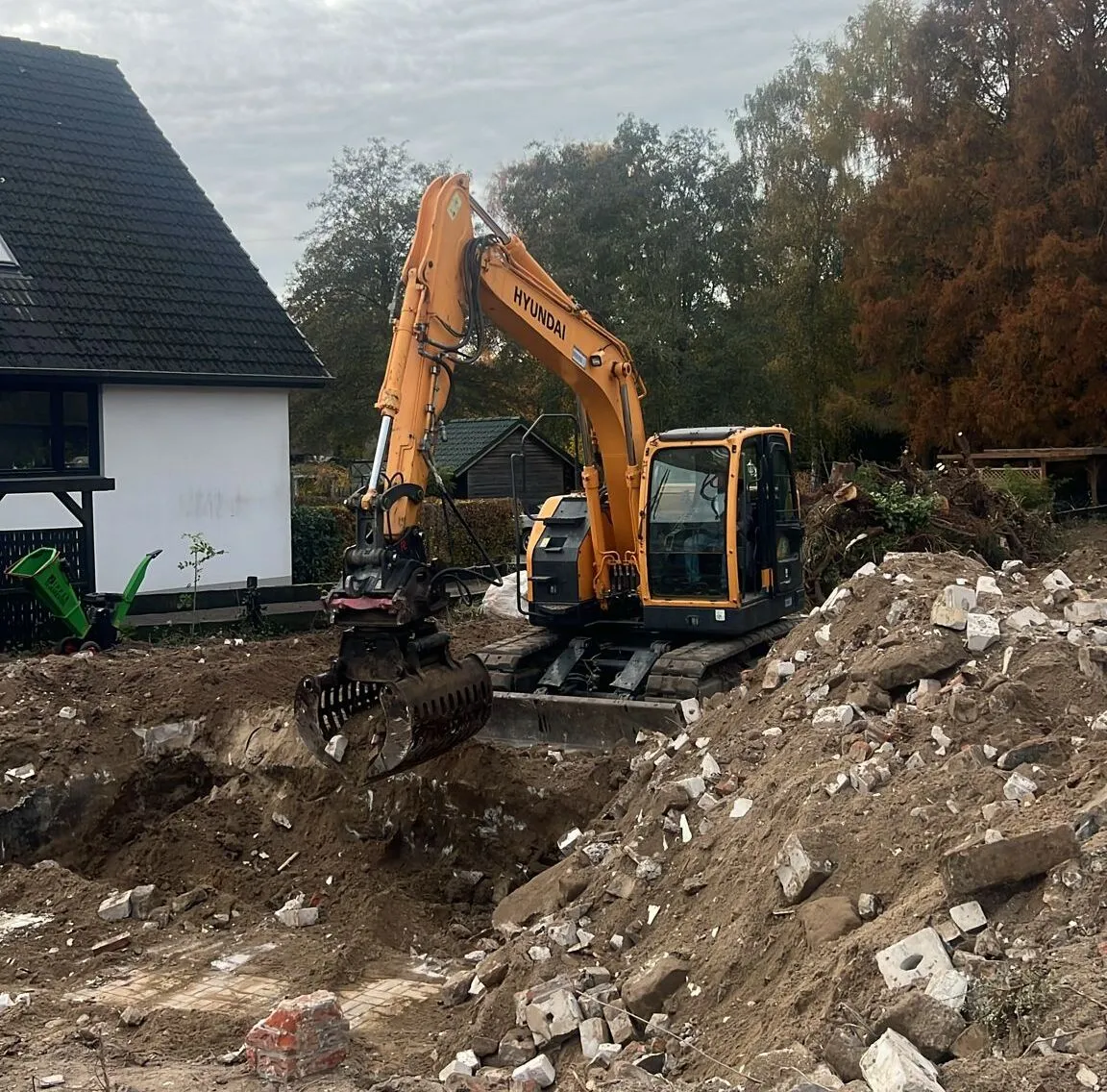 Ein gelber Hyundai-Bagger hebt Erde auf einer Baustelle neben einem Haus mit schwarzem Dach und weißen Wänden.