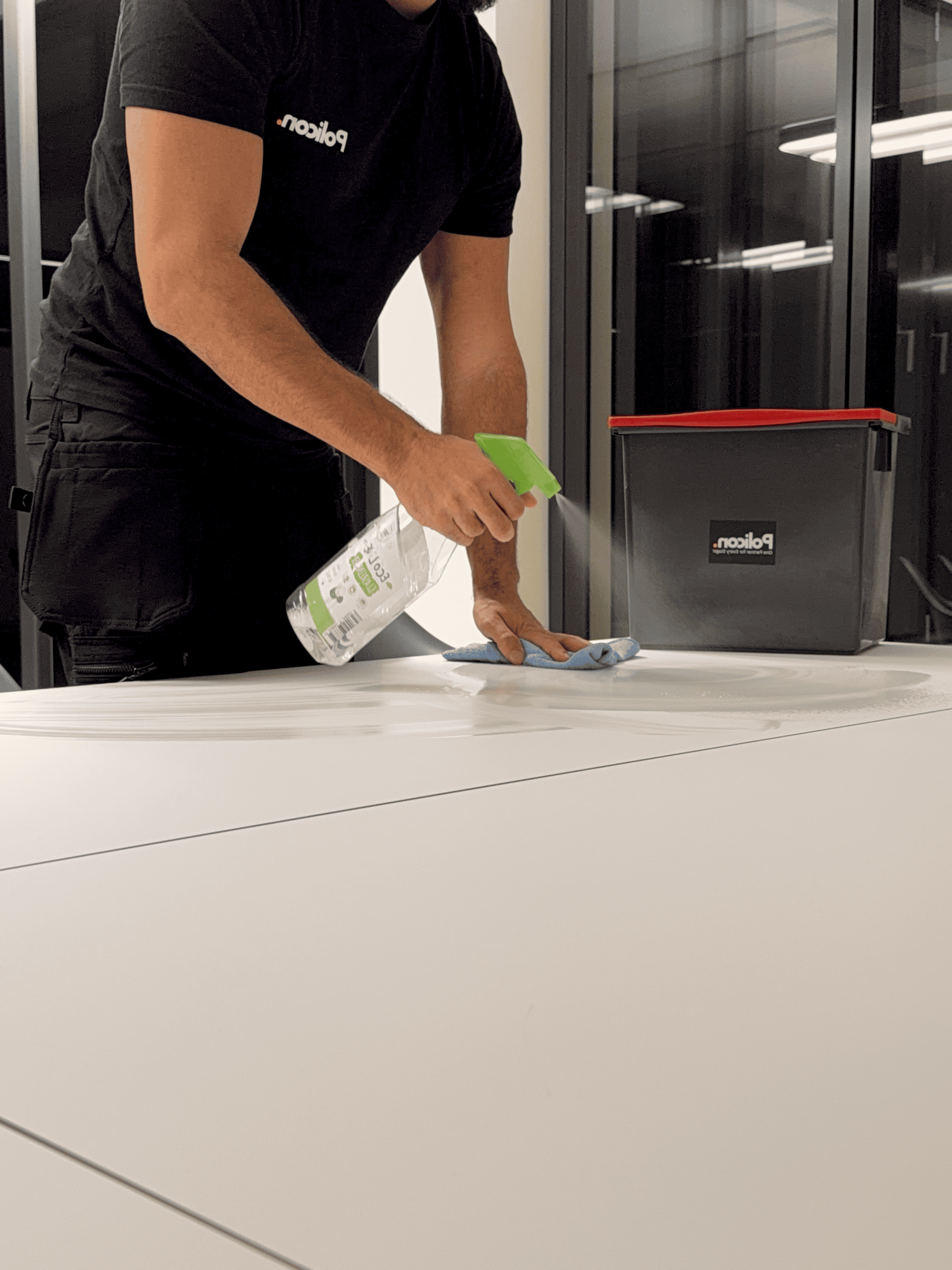 Person sprüht Reinigungsmittel auf eine weiße Oberfläche und wischt sie mit einem Tuch ab, ein Behälter mit rotem Deckel steht daneben.