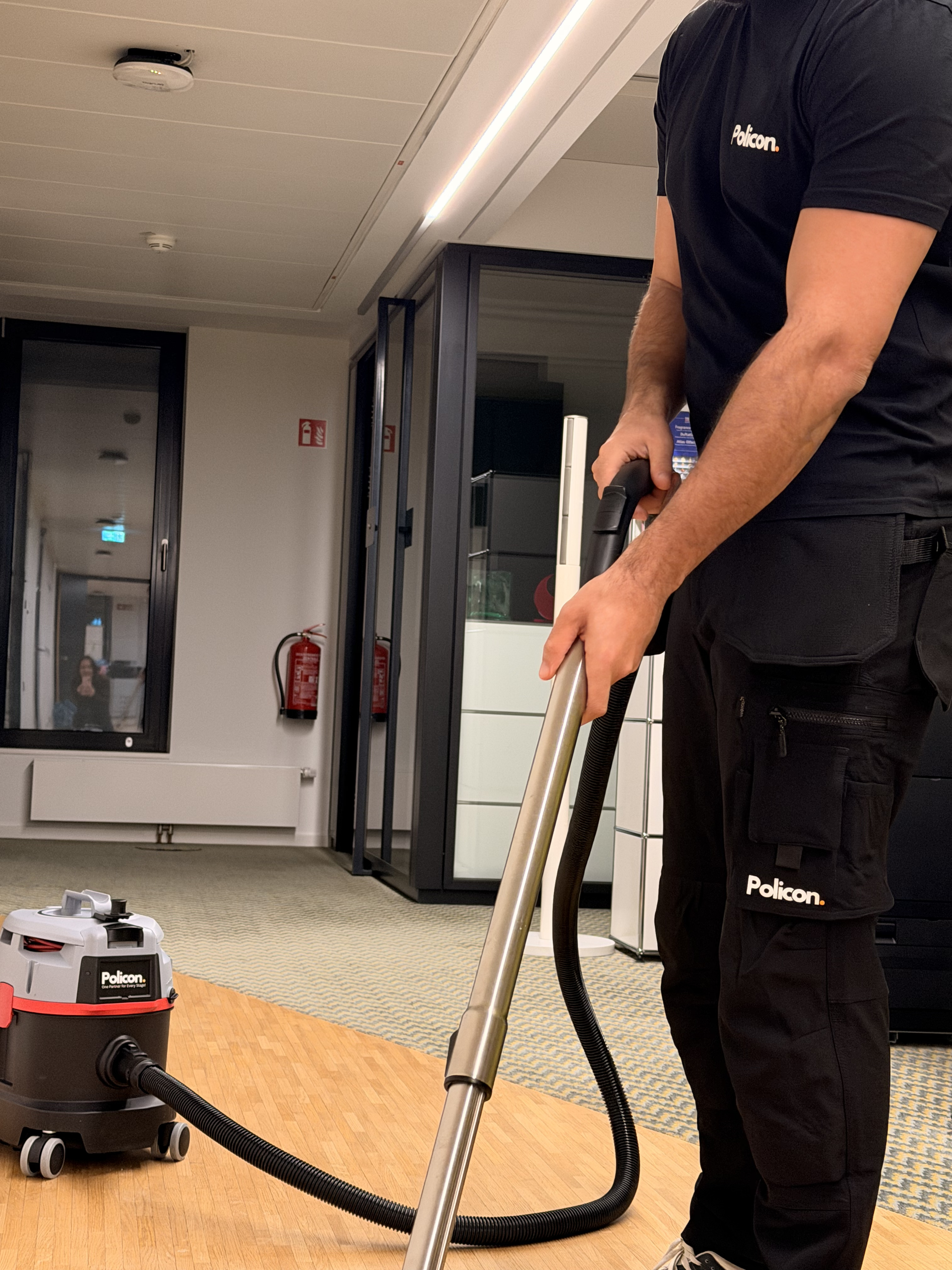 Person in schwarzer Policon-Arbeitskleidung saugt mit einem Staubsauger einen Boden im Büro.