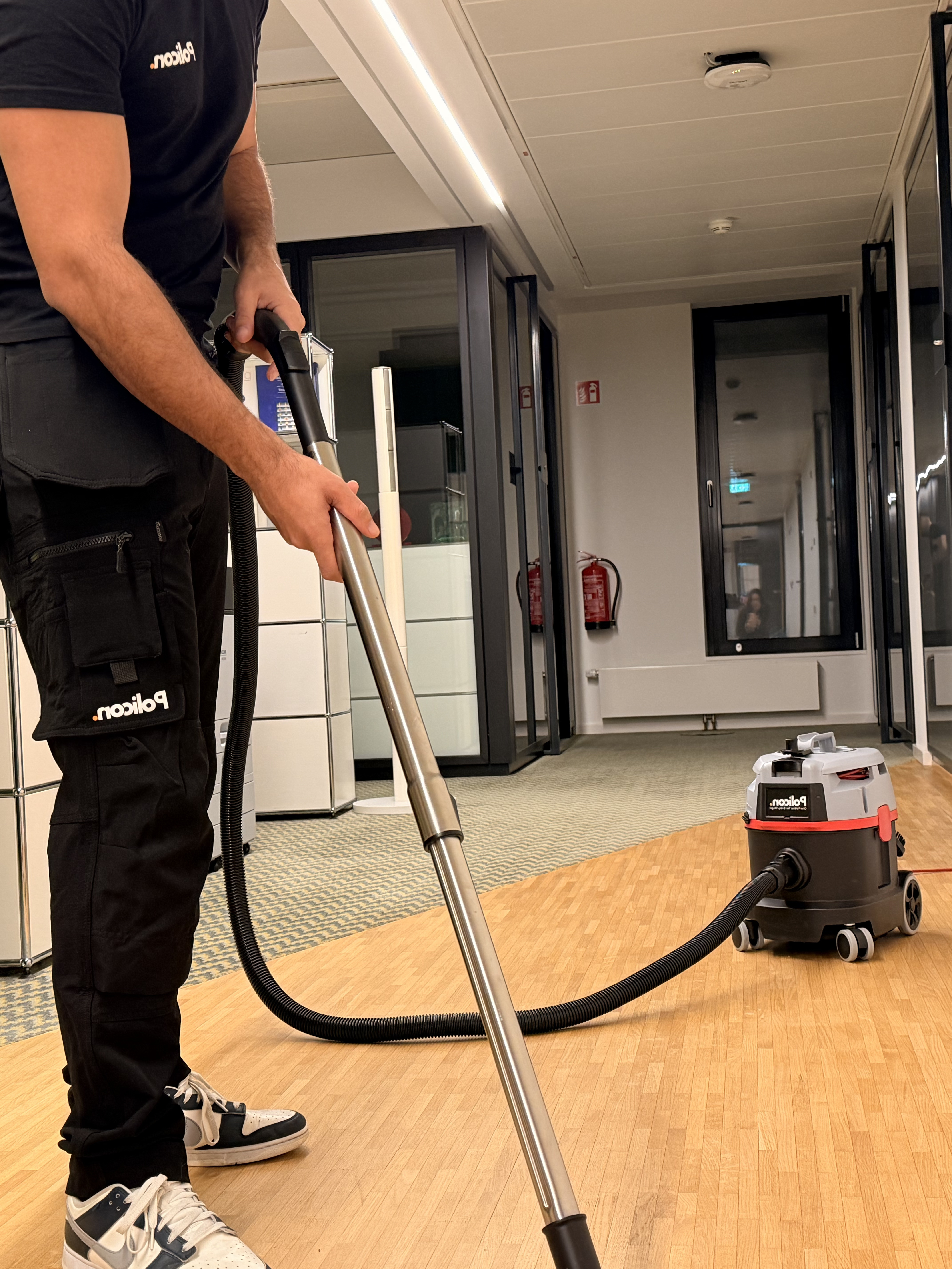 Mann in schwarzer Kleidung saugt mit einem Staubsauger einen Holzfußboden in einem modernen Büro.