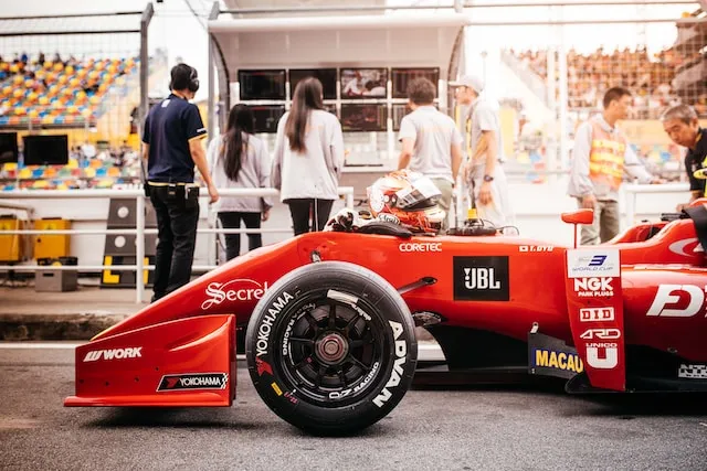Red Formula race car with branded tires and sponsors, parked on track with crew in background.