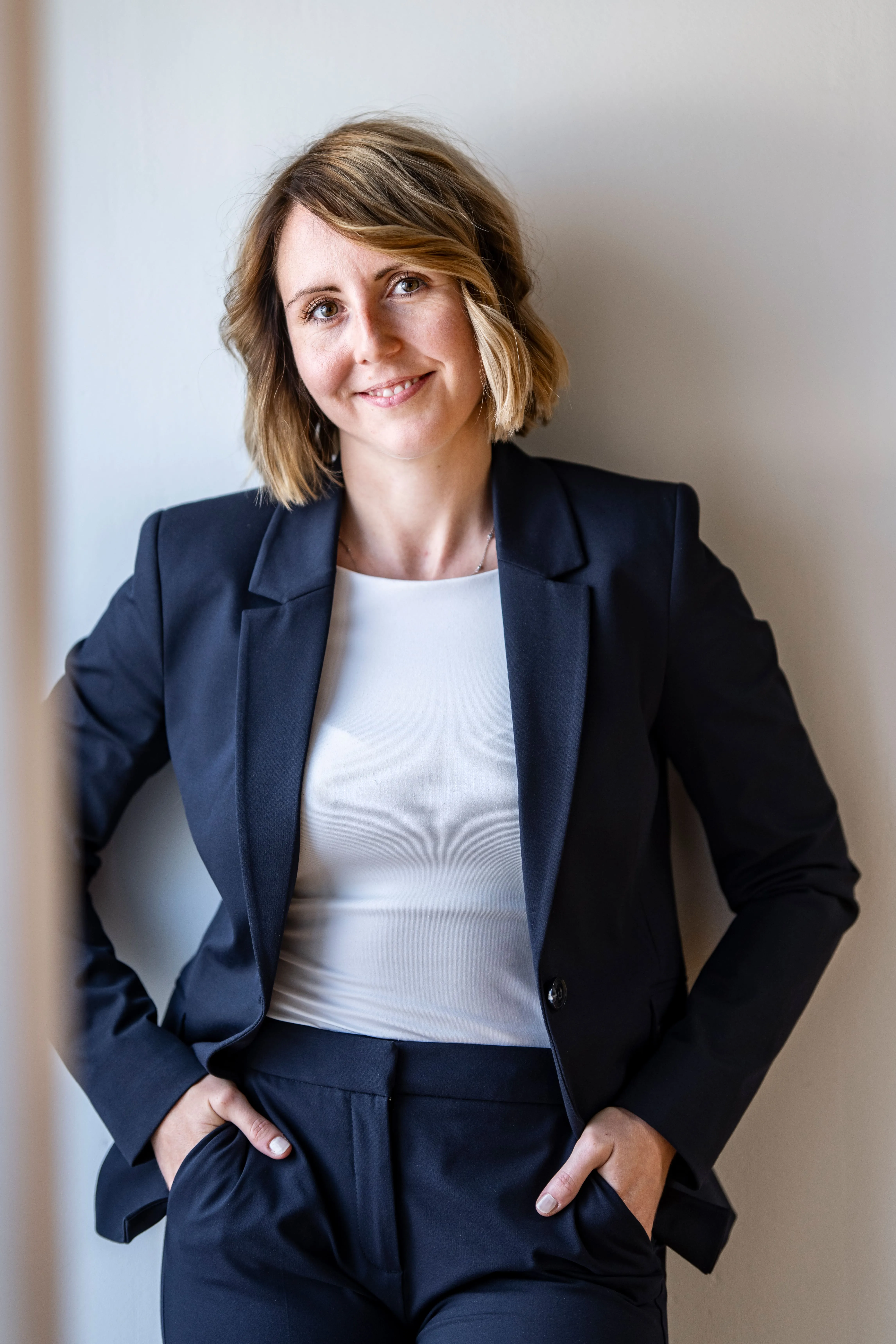 Stehende Frau im dunkelblauen Business-Outfit lehnt entspannt an einer Wand und lächelt.