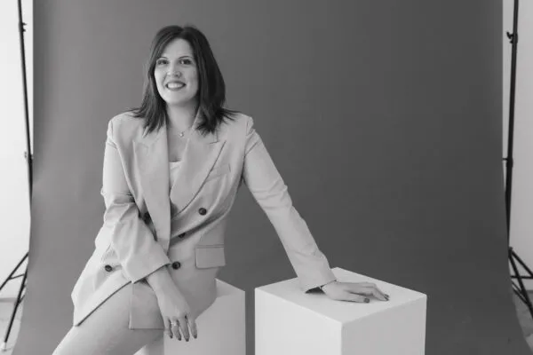 Porträt einer Frau im hellen Business-Outfit, sitzend und lächelnd vor neutralem Hintergrund.