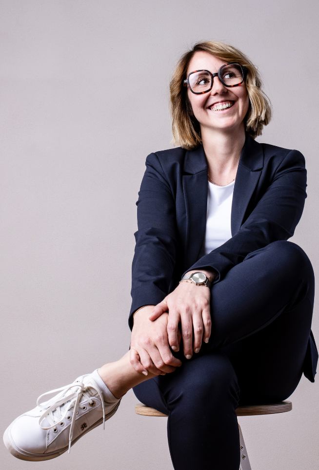 Rechtsanwältin im dunkelblauen Business-Outfit sitzt lächelnd auf einem Hocker vor neutralem Hintergrund.