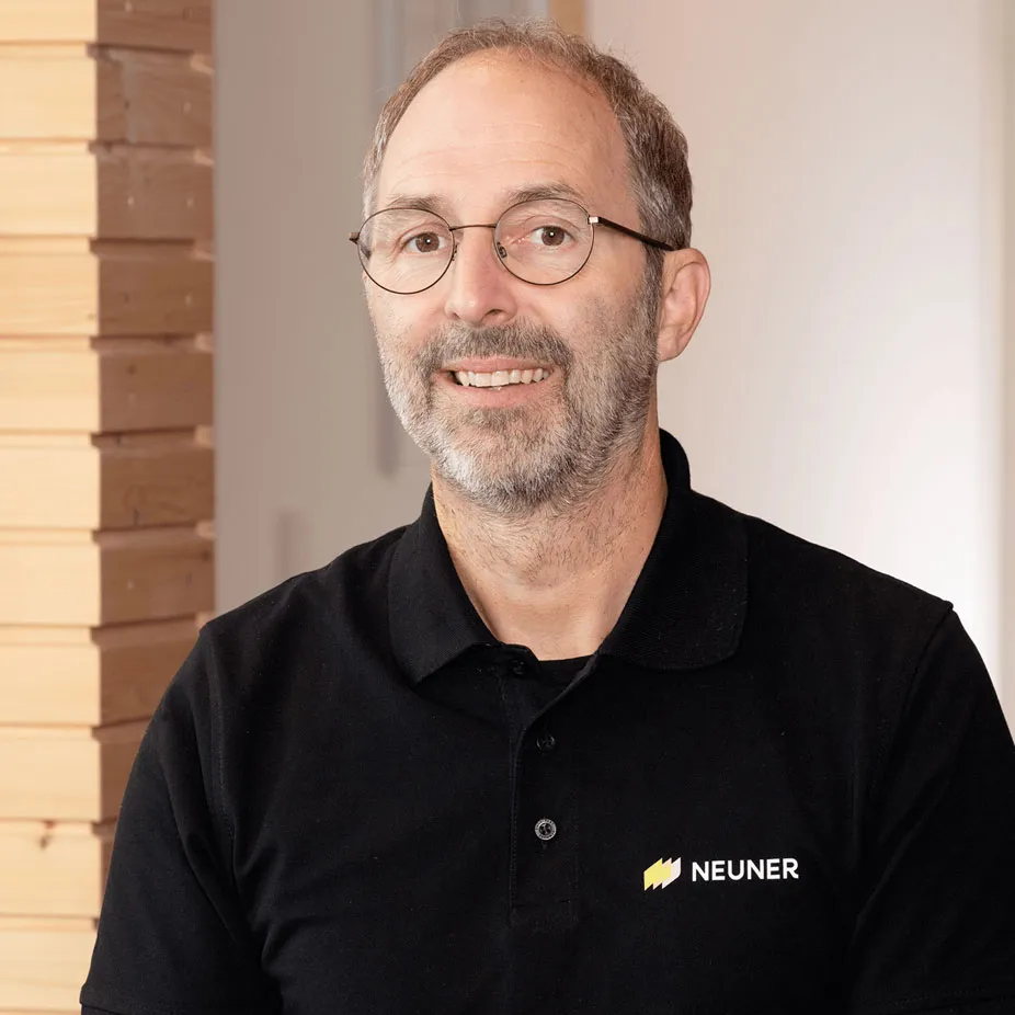 Portrait of a smiling NEUNER Insektenschutz team member in branded black polo shirt, standing indoors near stacked wood panels. Ideal for introducing company leadership or team values