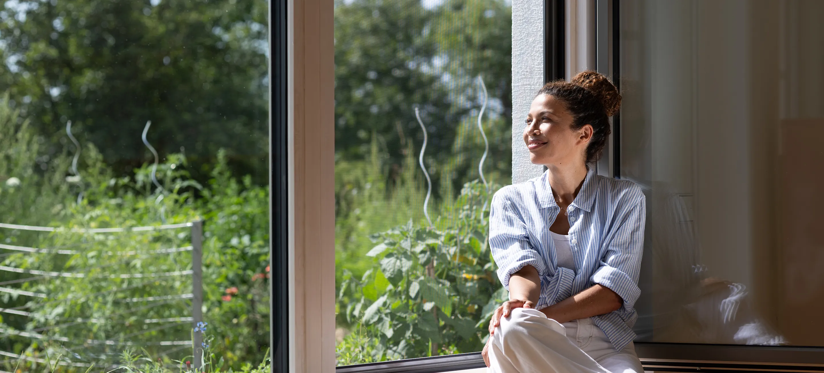 A smiling woman sits by a large window, basking in sunlight and looking outside. Lush greenery is visible in the background, conveying a serene mood