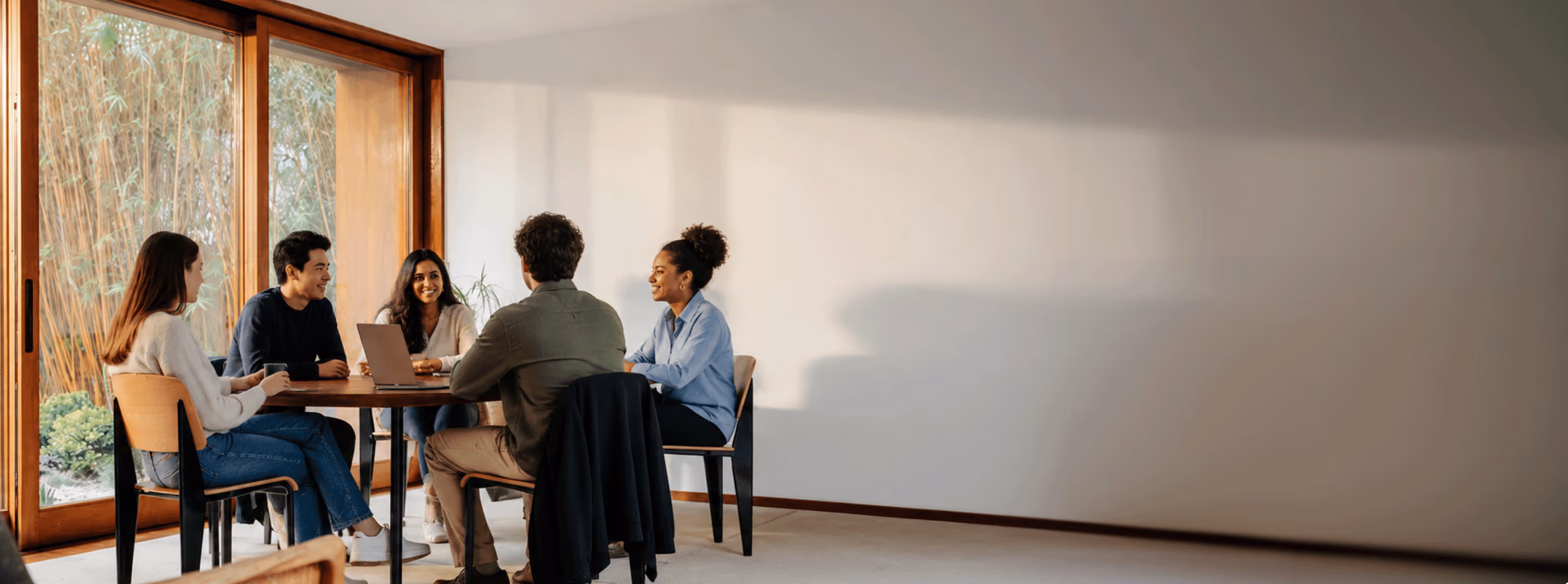 Five colleagues smiling and discussing a laptop screen in a sunny meeting room.