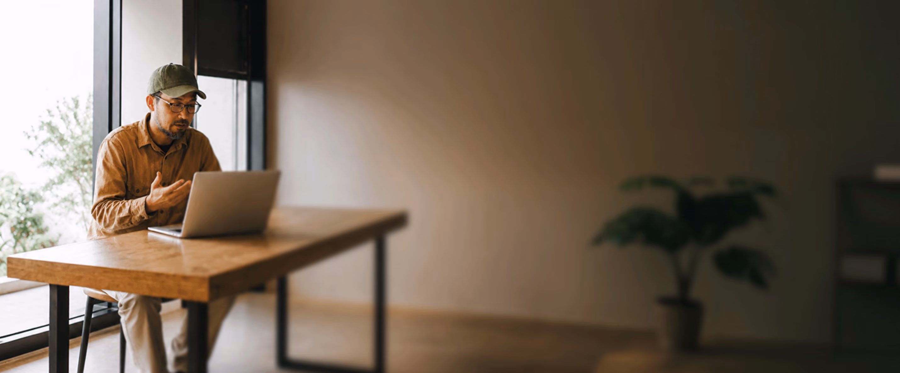 Man in a cap gesturing during a video call on his laptop at a wooden desk.