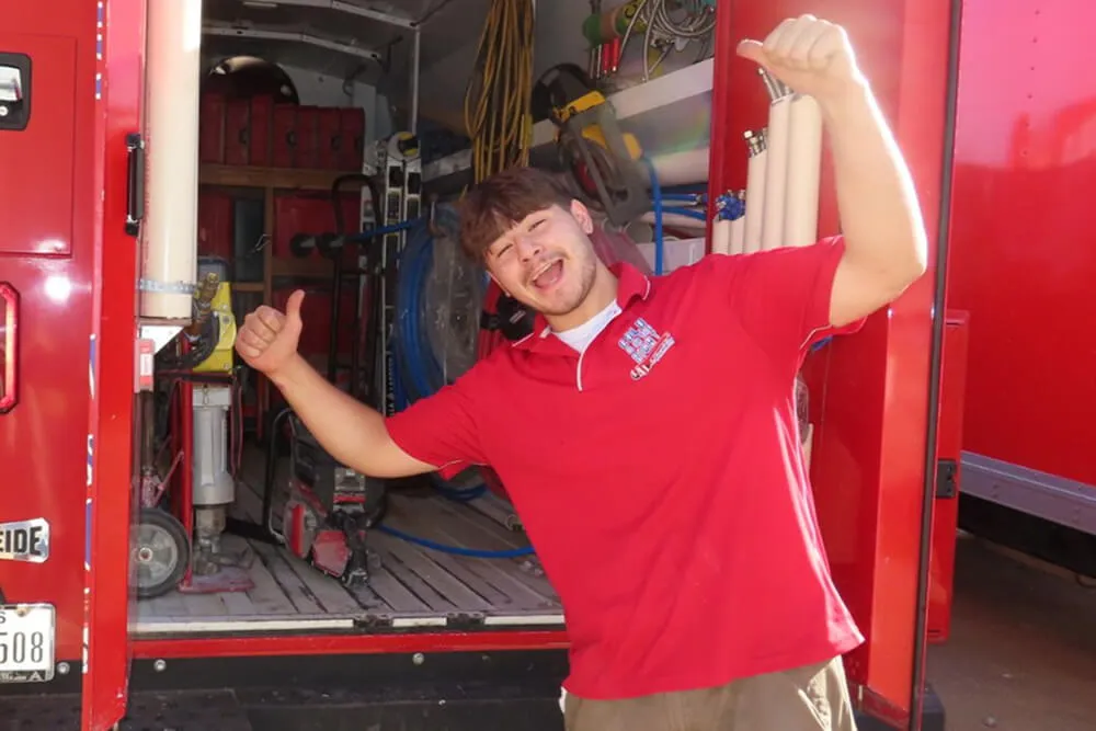 Smiling plumber in red shirt giving thumbs up in front of open work van.