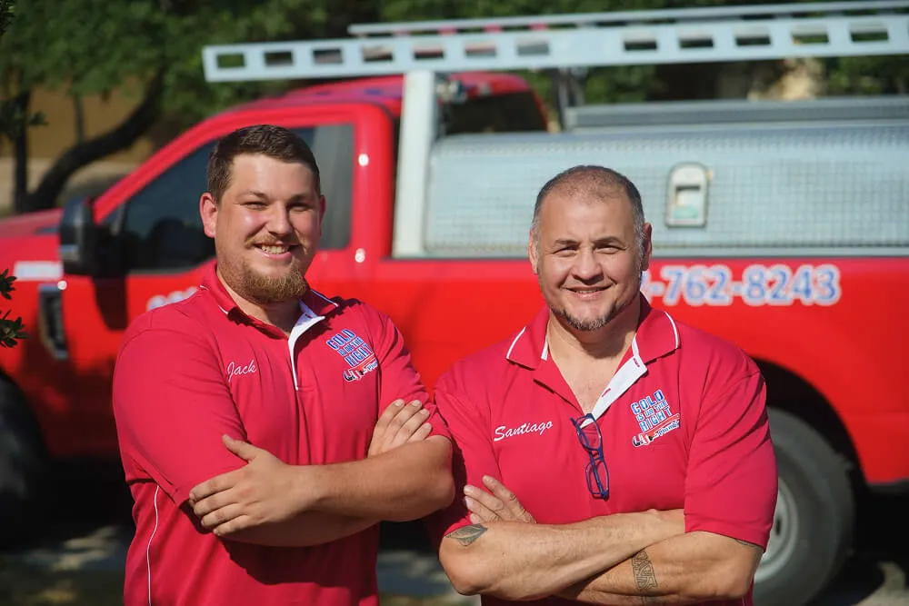 Two plumbers in red uniforms standing in front of red service truck.