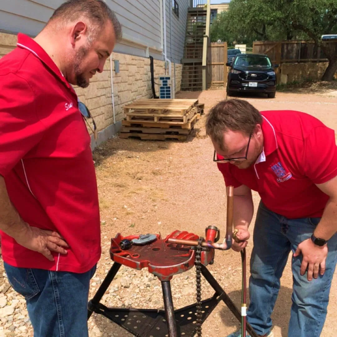 Two plumbers in red shirts work on copper pipe outdoors.