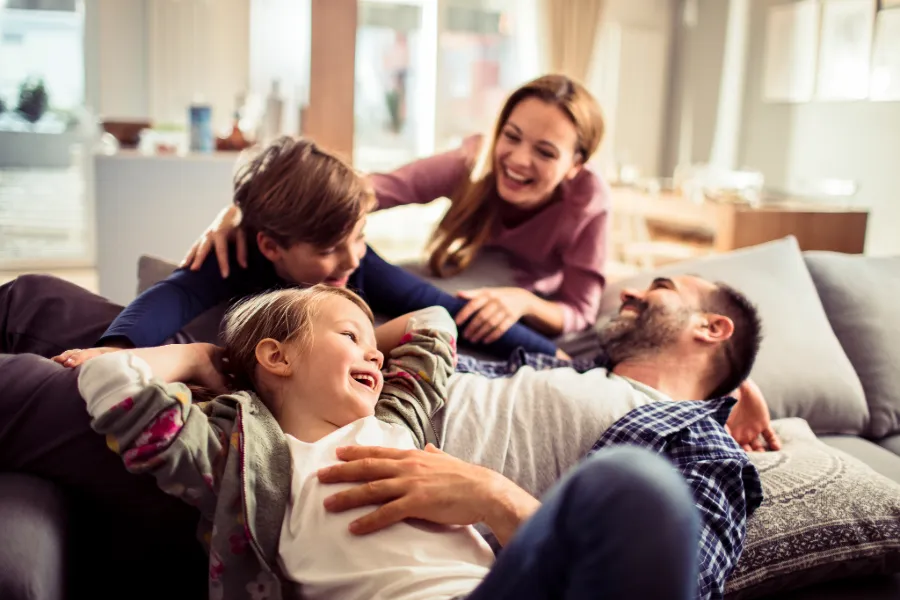 A family of four laughing and relaxing together on a couch in a bright living room.