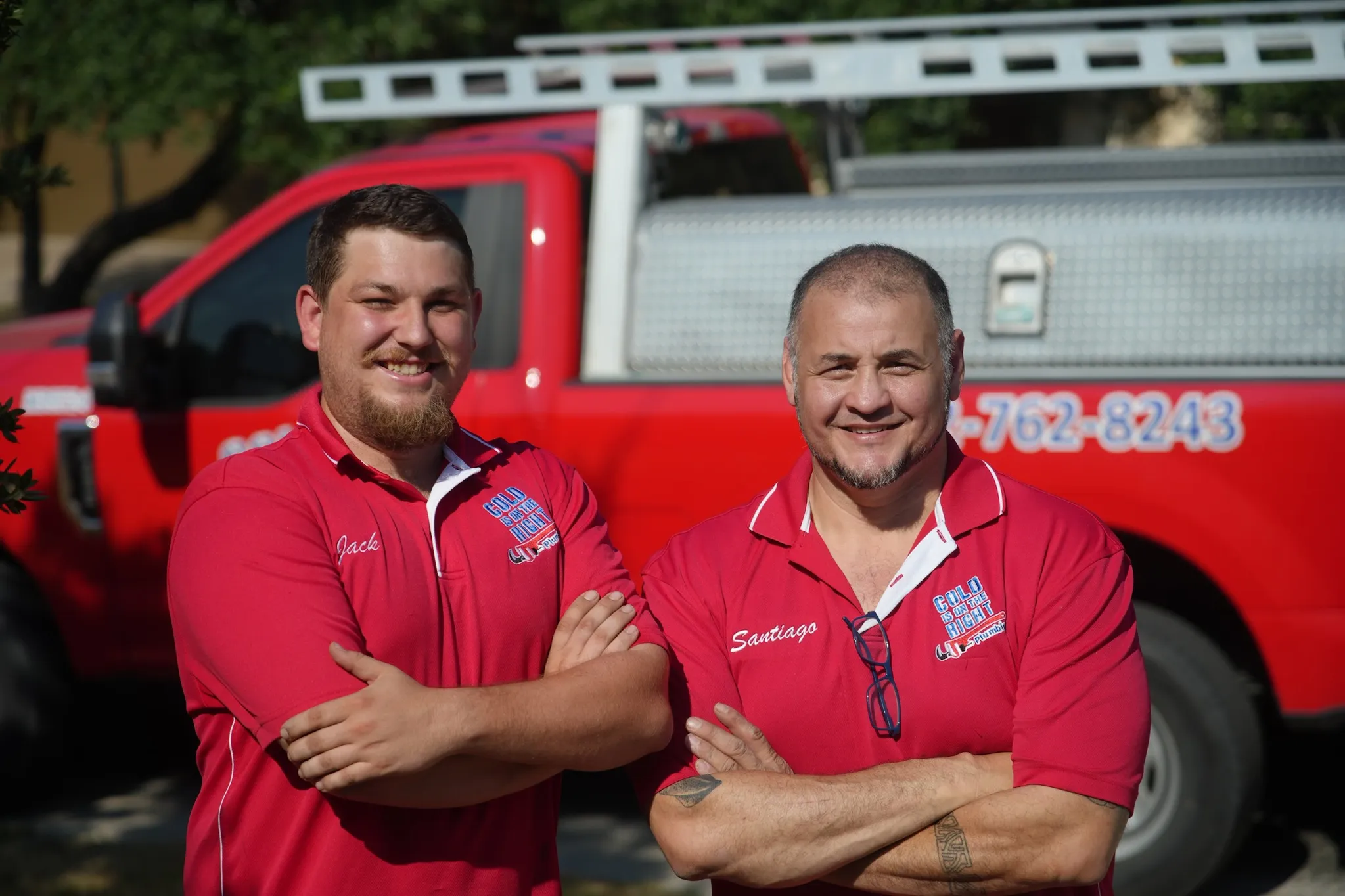 Two smiling HVAC technicians wearing red shirts with 'Cold is on the Right' logos standing in front of a red service truck with folded ladder.