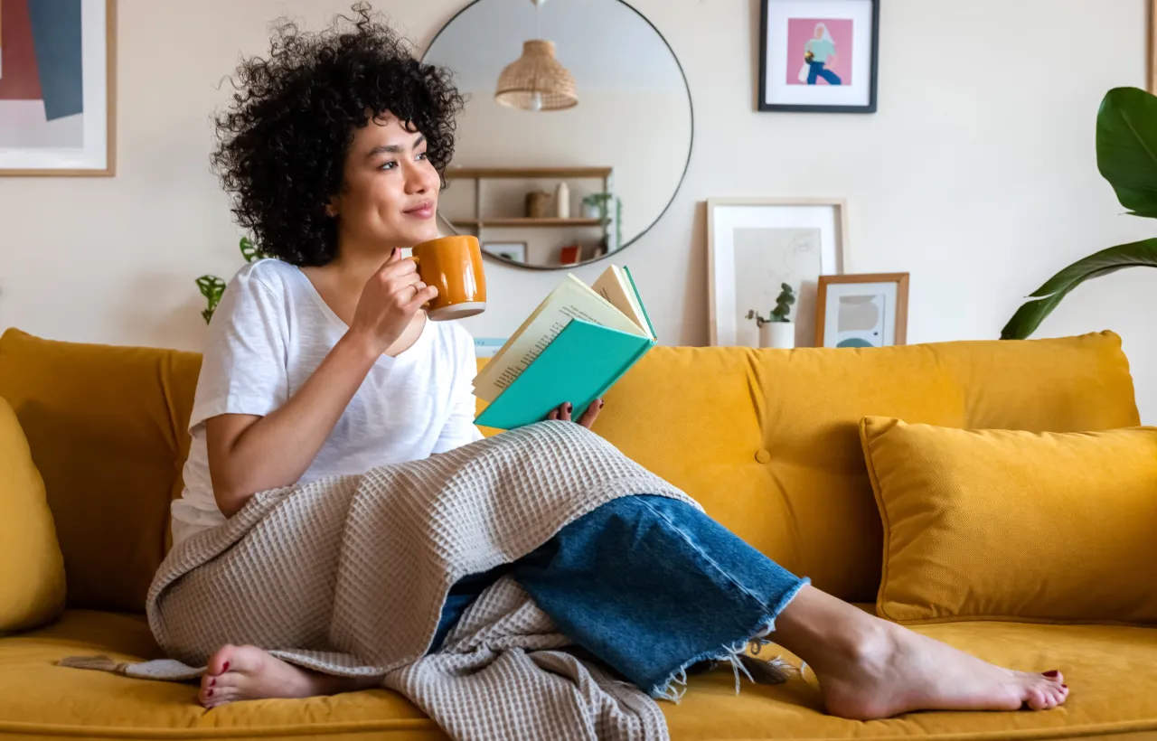 Woman sitting on a yellow sofa wrapped in a blanket, holding an open book and drinking from an orange mug.