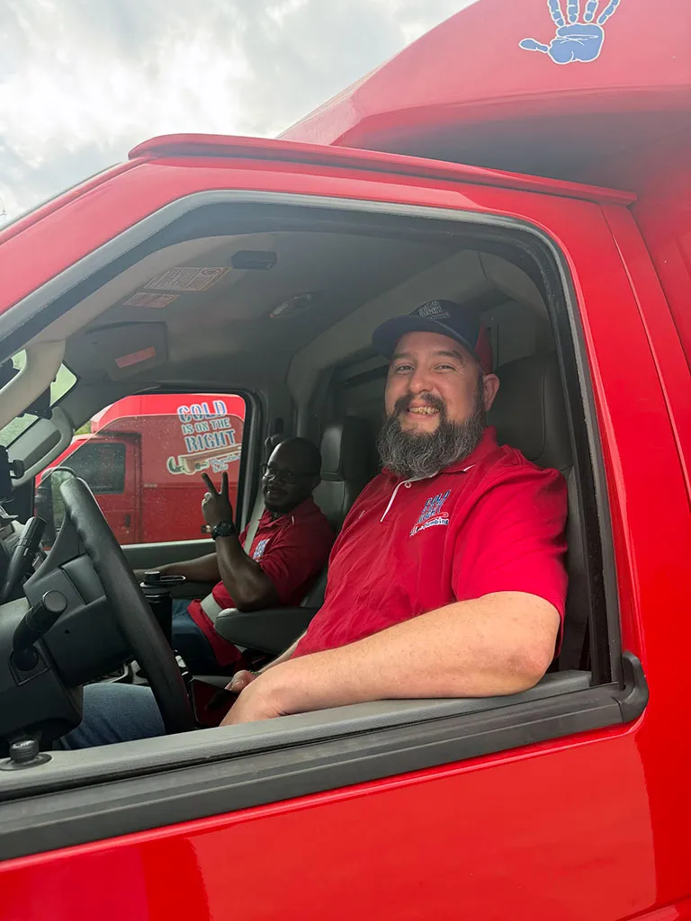 Two men in red shirts and caps sitting inside a red delivery truck, one smiling at the camera and the other showing a peace sign.