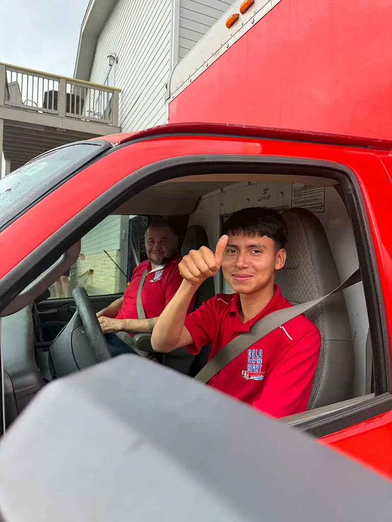 Two men in red shirts sitting inside a red delivery truck, one smiling and giving a thumbs-up from the driver's seat.